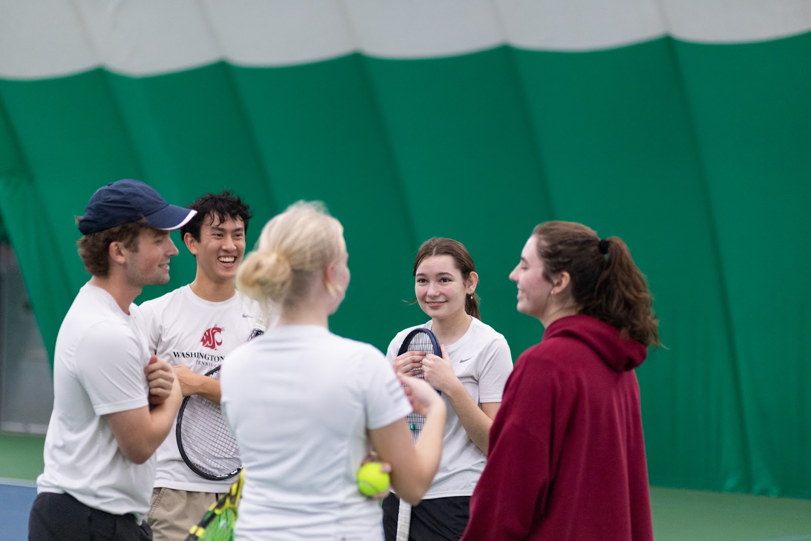 Group of young adults with tennis racquets smiling and talking on an indoor tennis court.