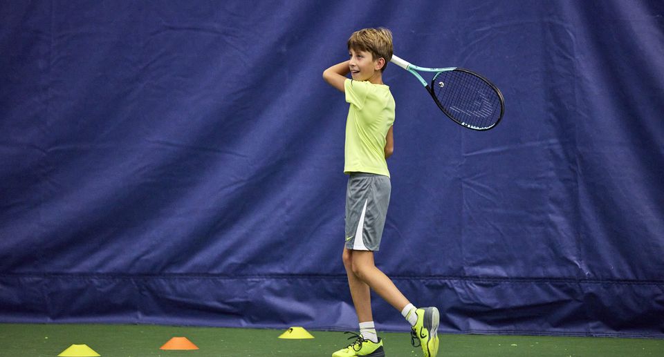 A young boy in a yellow shirt and gray shorts swings a tennis racket indoors, smiling, with colored cones on the court.