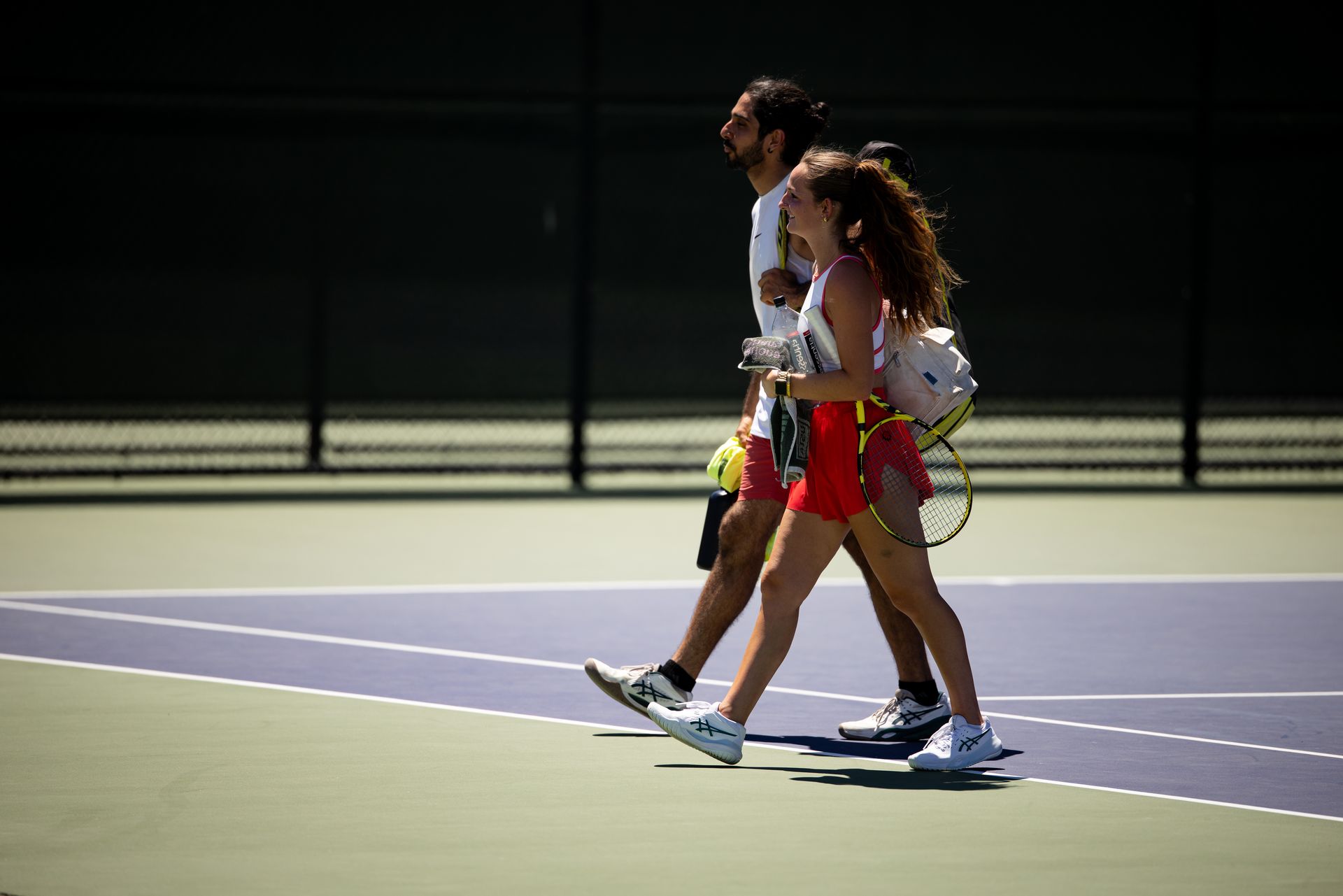 Two people walking off a tennis court with rackets and water bottles, smiling under bright sunlight.