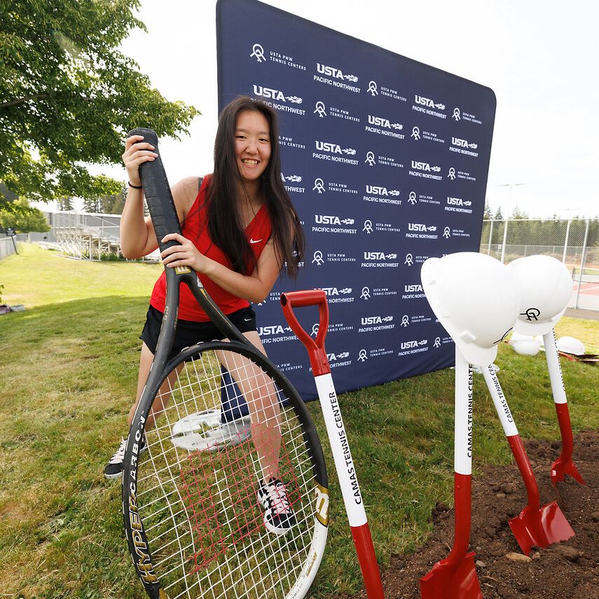 Smiling woman holding a giant tennis racket next to red shovels at an outdoor event, with a branded backdrop in the background.