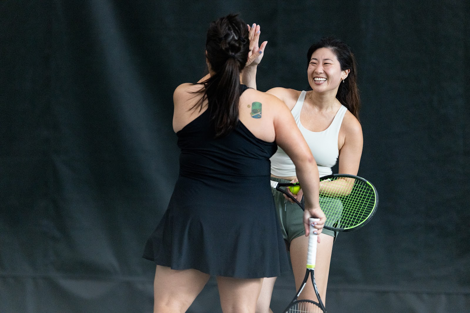 Two women on an indoor tennis court joyfully high-five each other, both holding rackets and smiling widely.