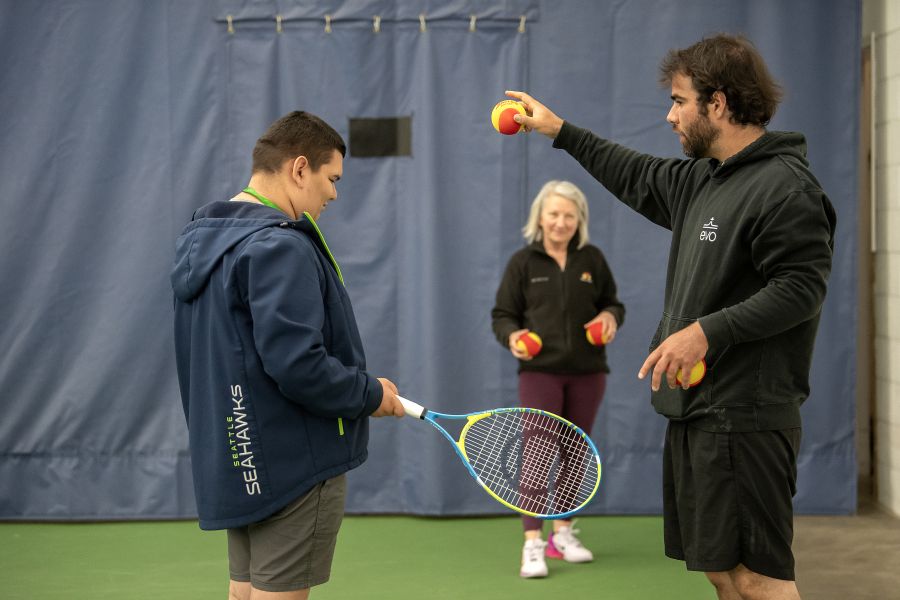 A coach instructs a young man with a tennis racket while a smiling woman holds tennis balls in the background on an indoor court.