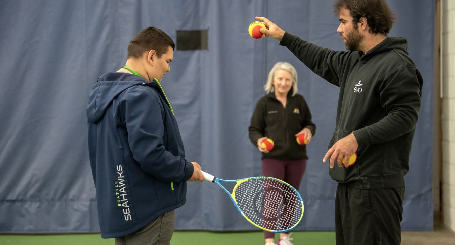 A coach instructs a young man with a tennis racket while a smiling woman holds tennis balls in the background on an indoor court.