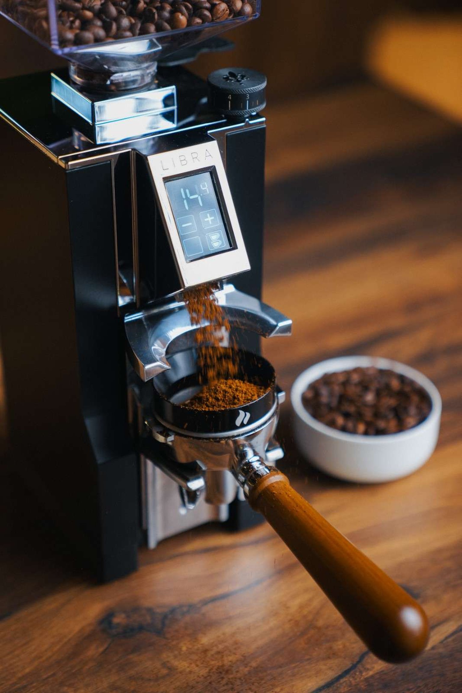 Espresso machine grinding coffee beans into a portafilter. Digital display shows settings. Bowl of whole beans in the background.