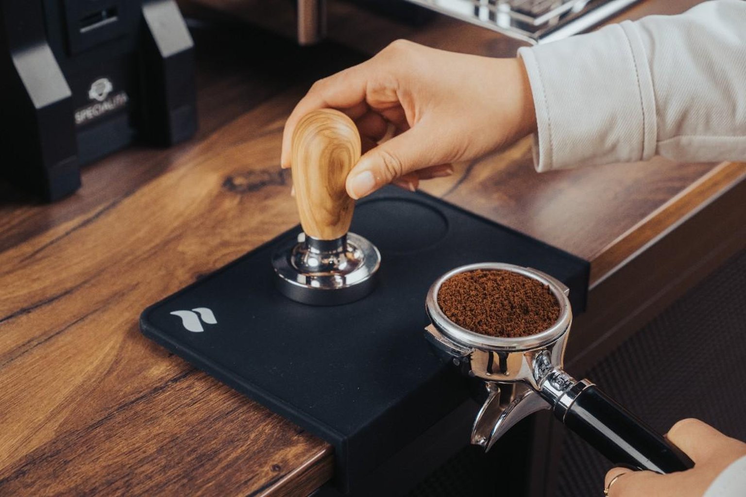 A hand uses a wooden-handled tamper on coffee grounds in a portafilter on a wooden countertop.