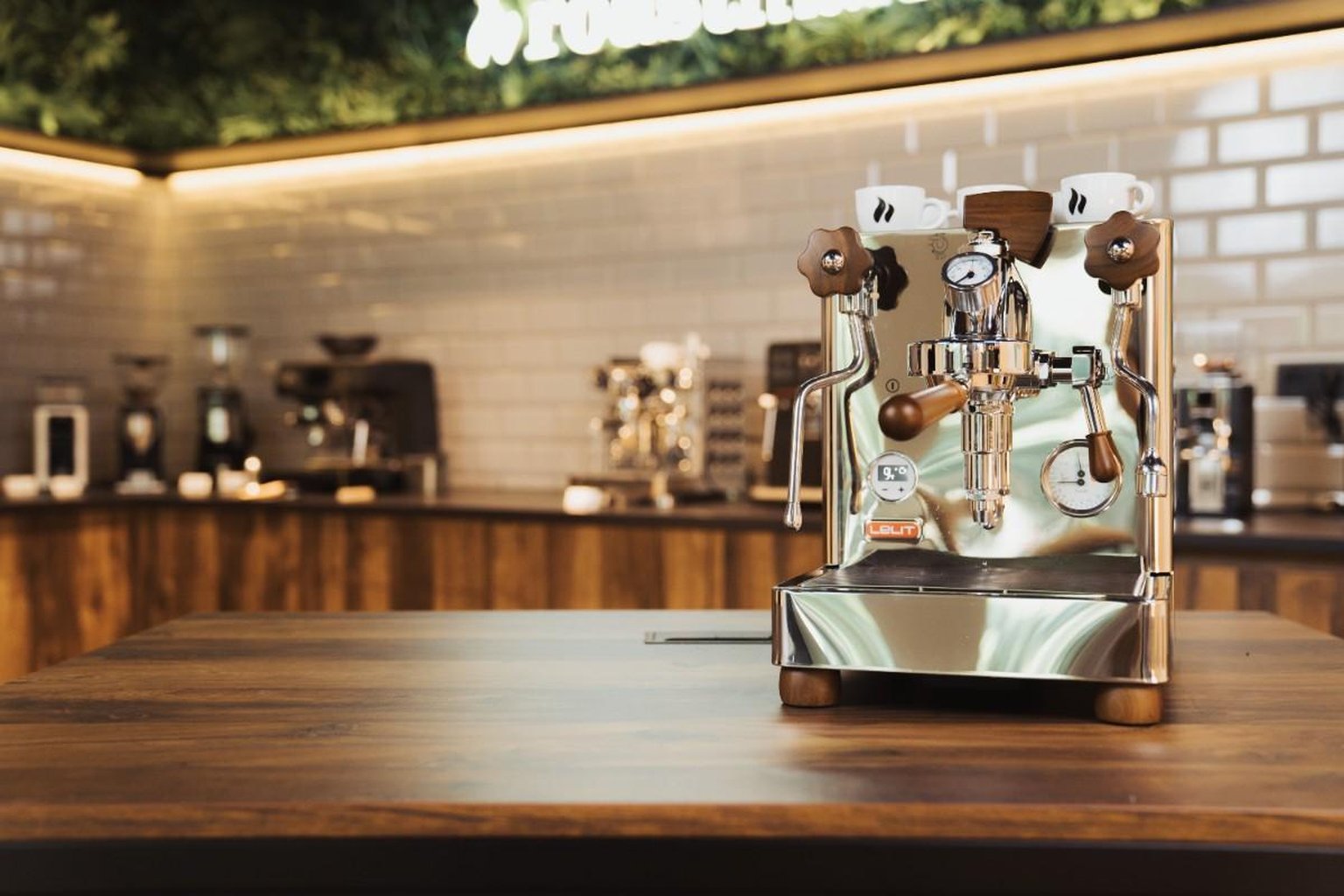 Shiny espresso machine with wooden knobs on a wooden counter, against a tiled wall in a modern cafe setting.