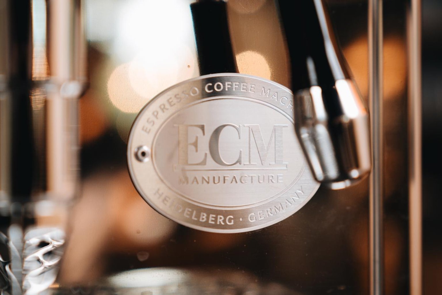 Close-up of a silver ECM Manufacture logo on an espresso machine, with blurred background lighting.