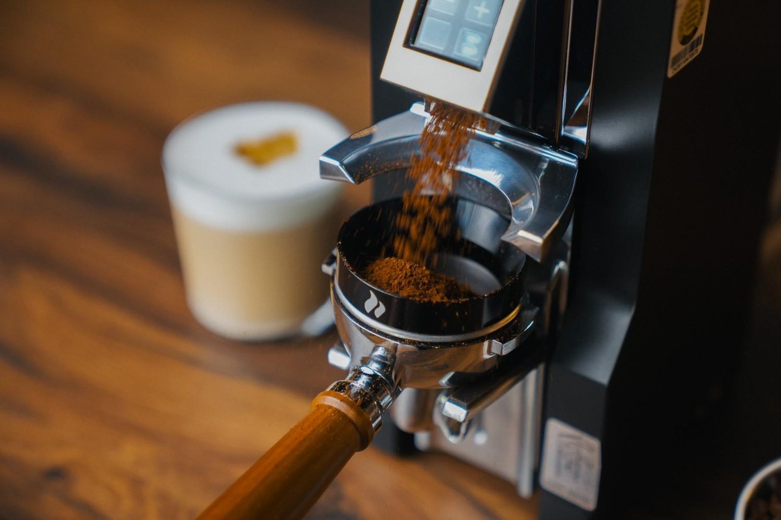 Coffee grinder dispensing grounds into a portafilter, with a cup of cappuccino on a wooden table in the background.