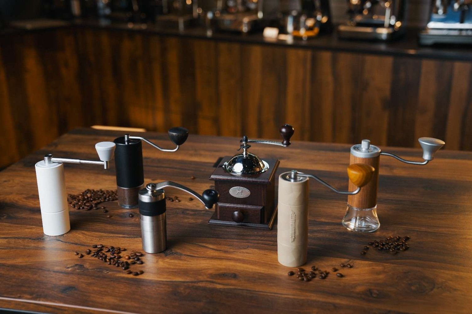A variety of manual coffee grinders displayed on a wooden table, surrounded by scattered coffee beans, with a vintage grinder in the center.