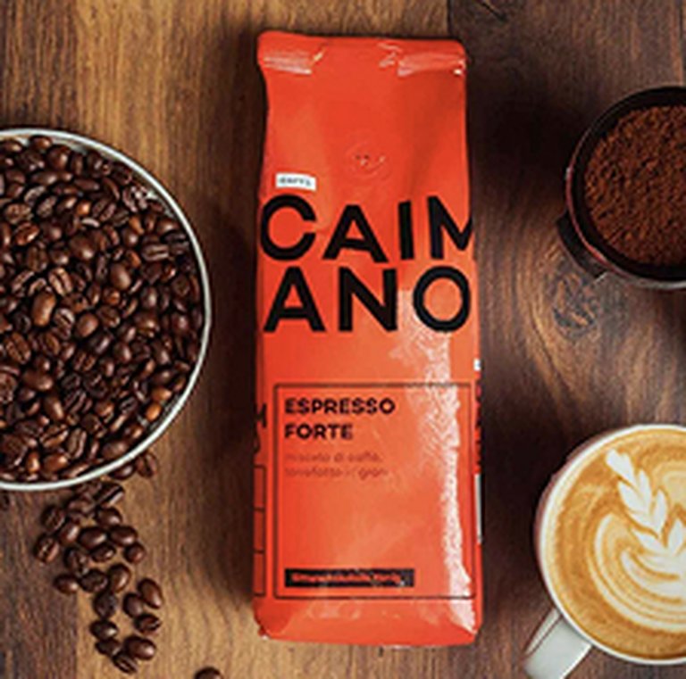 Bag of Caimano espresso forte on a wooden table with a bowl of coffee beans, ground coffee, and a cup of latte with latte art.
