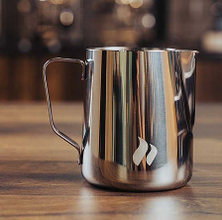 Stainless steel milk frothing pitcher with a handle, on a wooden surface, soft-focus background.
