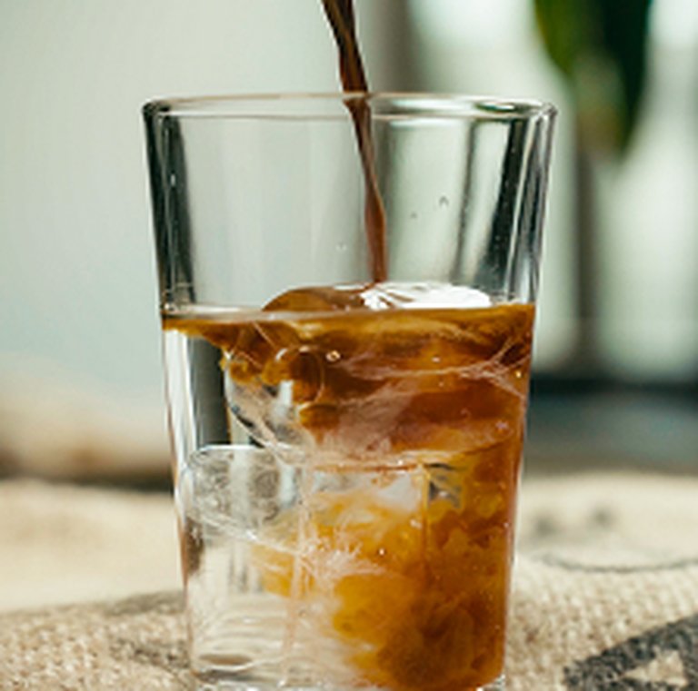 Iced coffee being poured into a glass filled with ice, set on a textured surface with a blurred background.