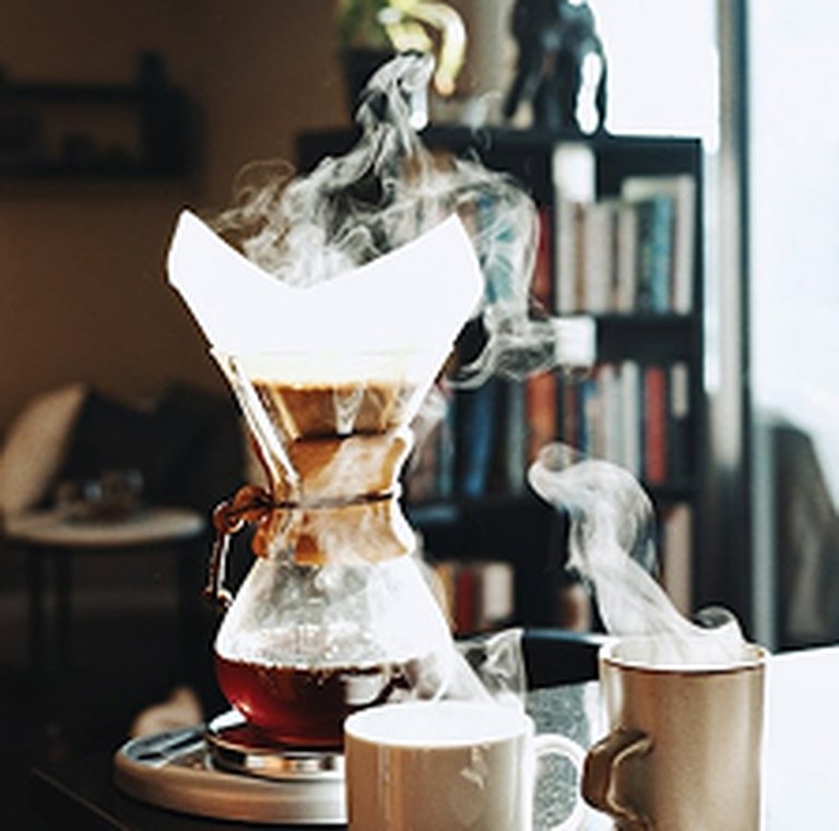 A Chemex coffee maker brewing with steam rising, next to steaming mugs on a table in a cozy room with books and soft lighting.