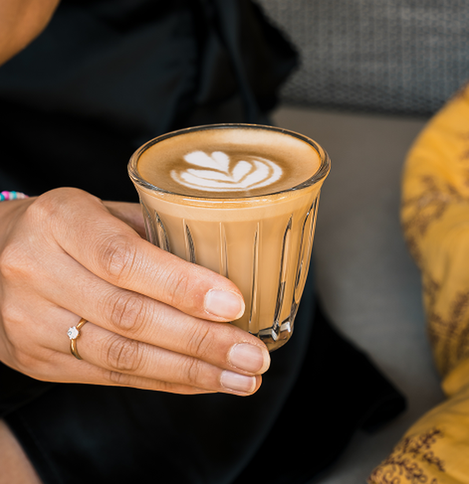 Close-up of a hand holding a clear, ribbed glass of latte with elegant heart-shaped latte art. A subtle ring adorns a finger.