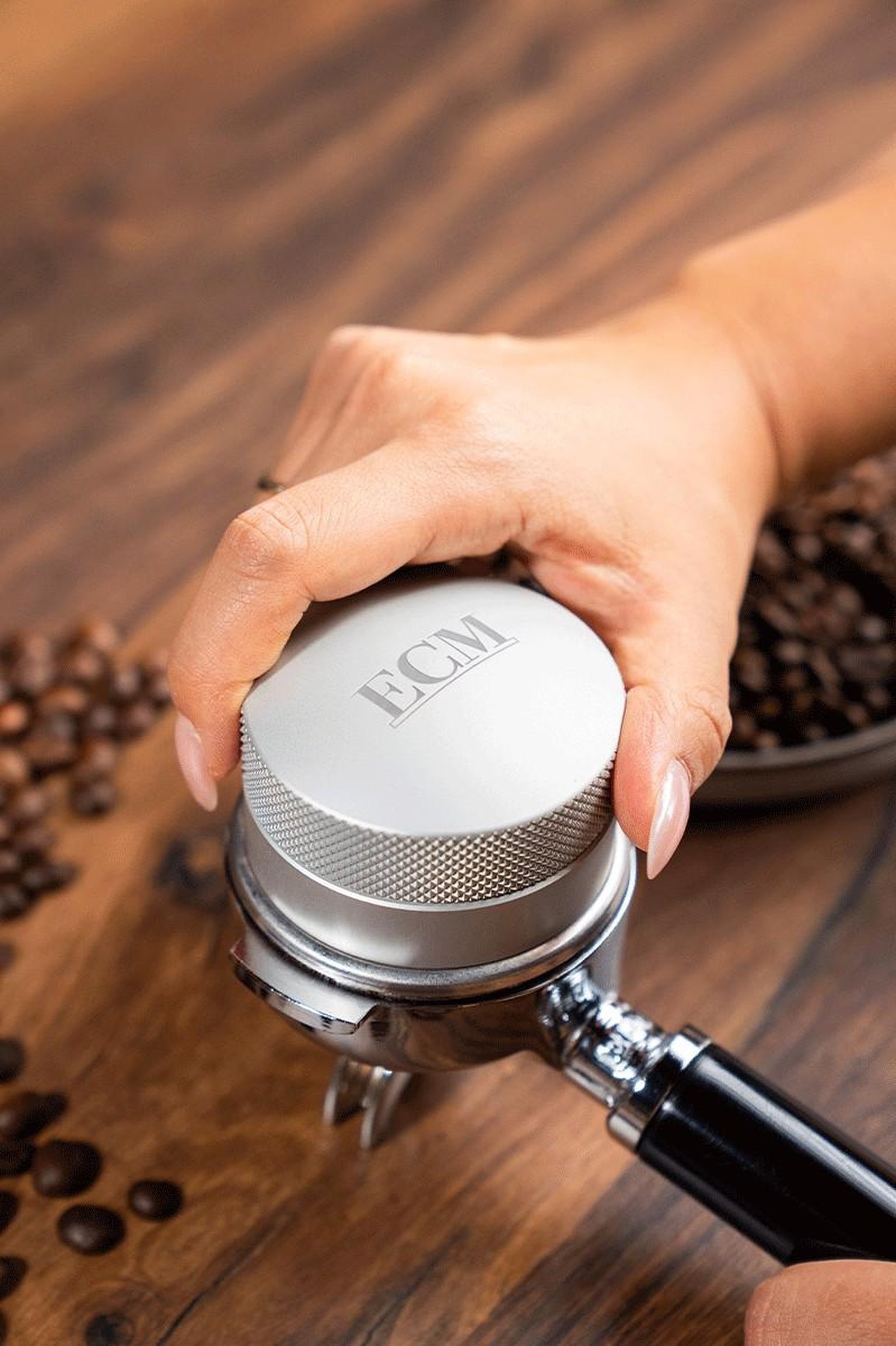 Hand using a silver espresso tamper on a portafilter, with coffee beans on a wooden surface.