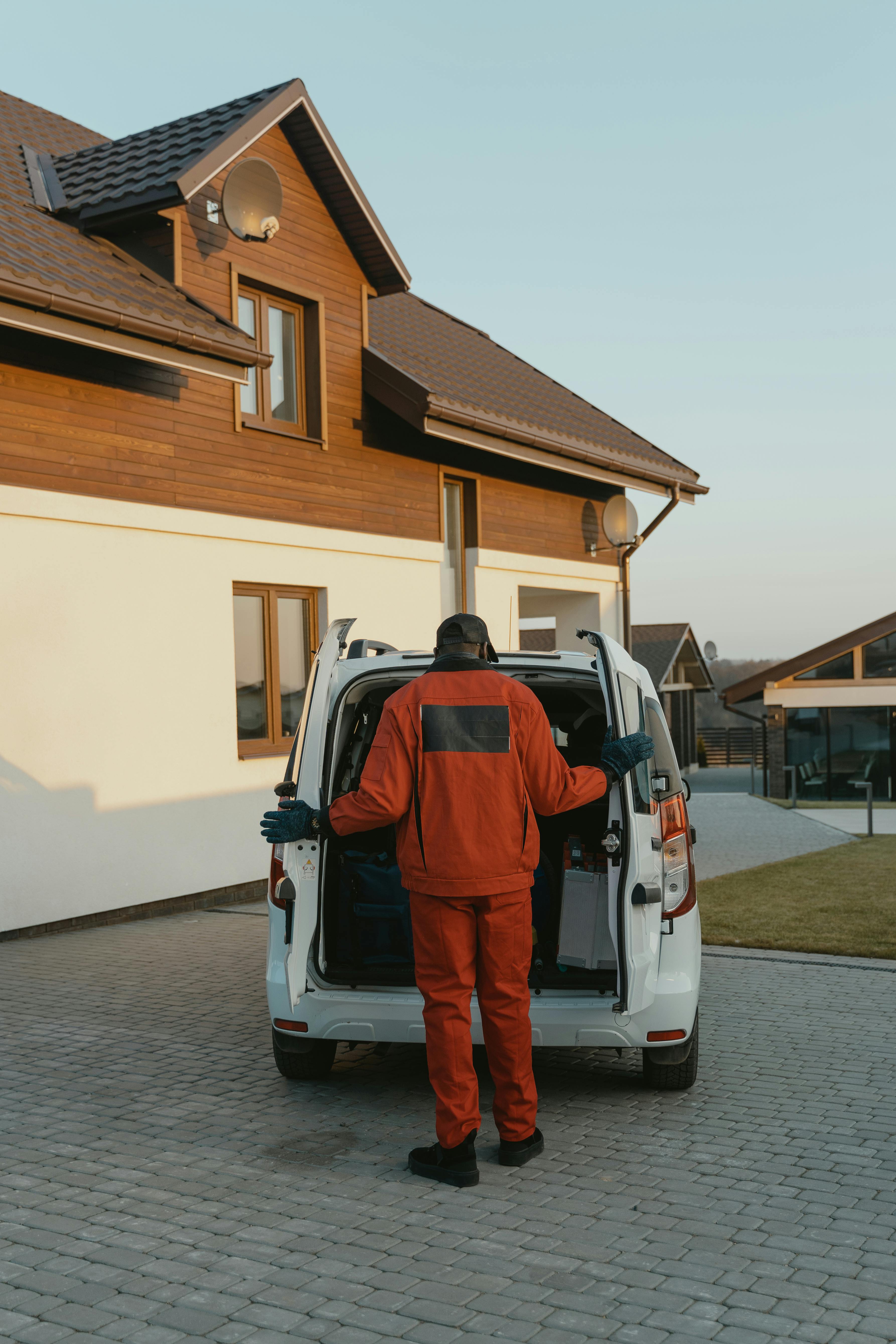 A person in a red uniform unloads items from a white van parked on a cobblestone driveway, in front of a modern house with wood siding.