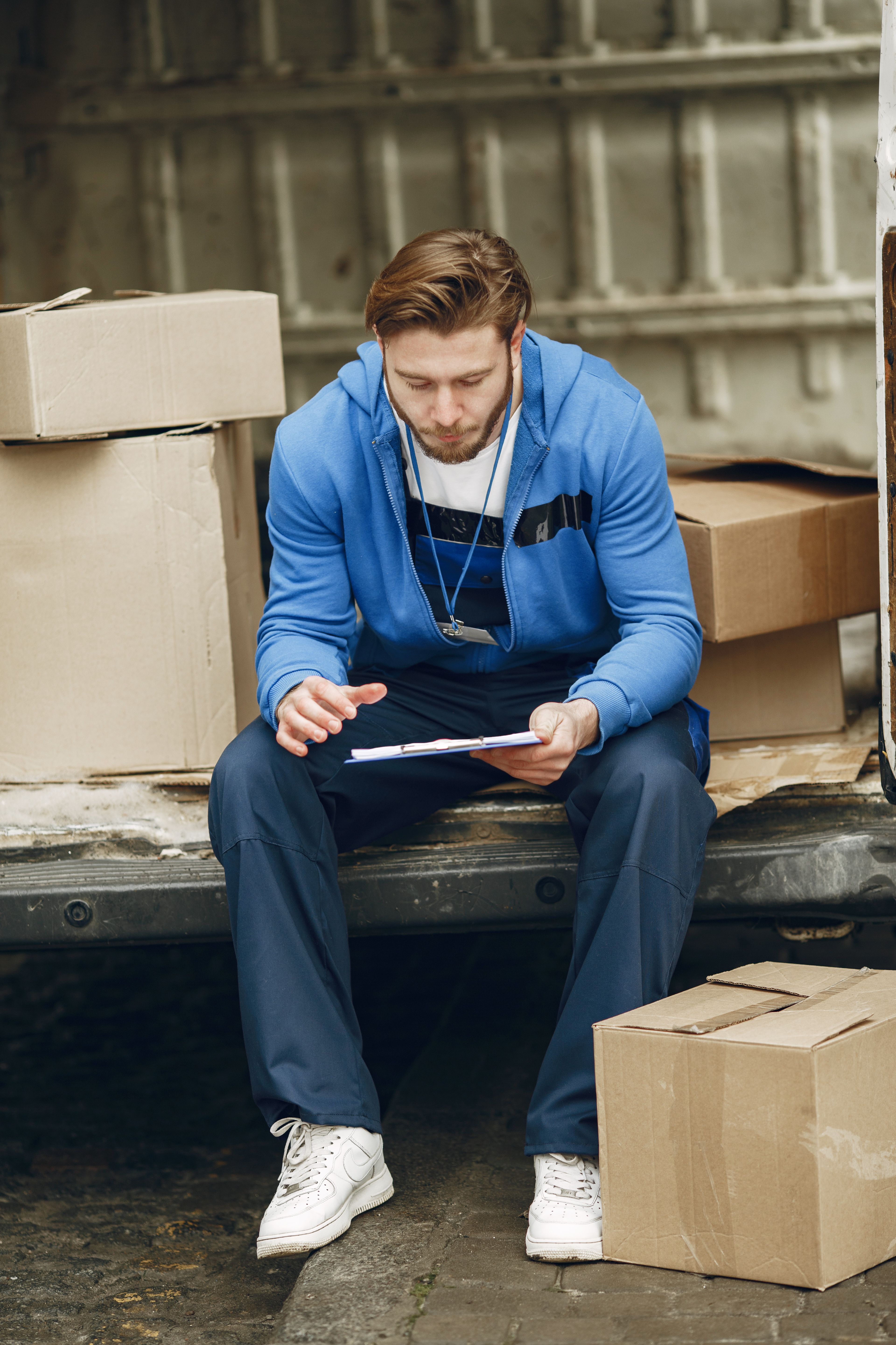 Young man in blue hoodie sitting in a van, reading a clipboard with cardboard boxes around him.