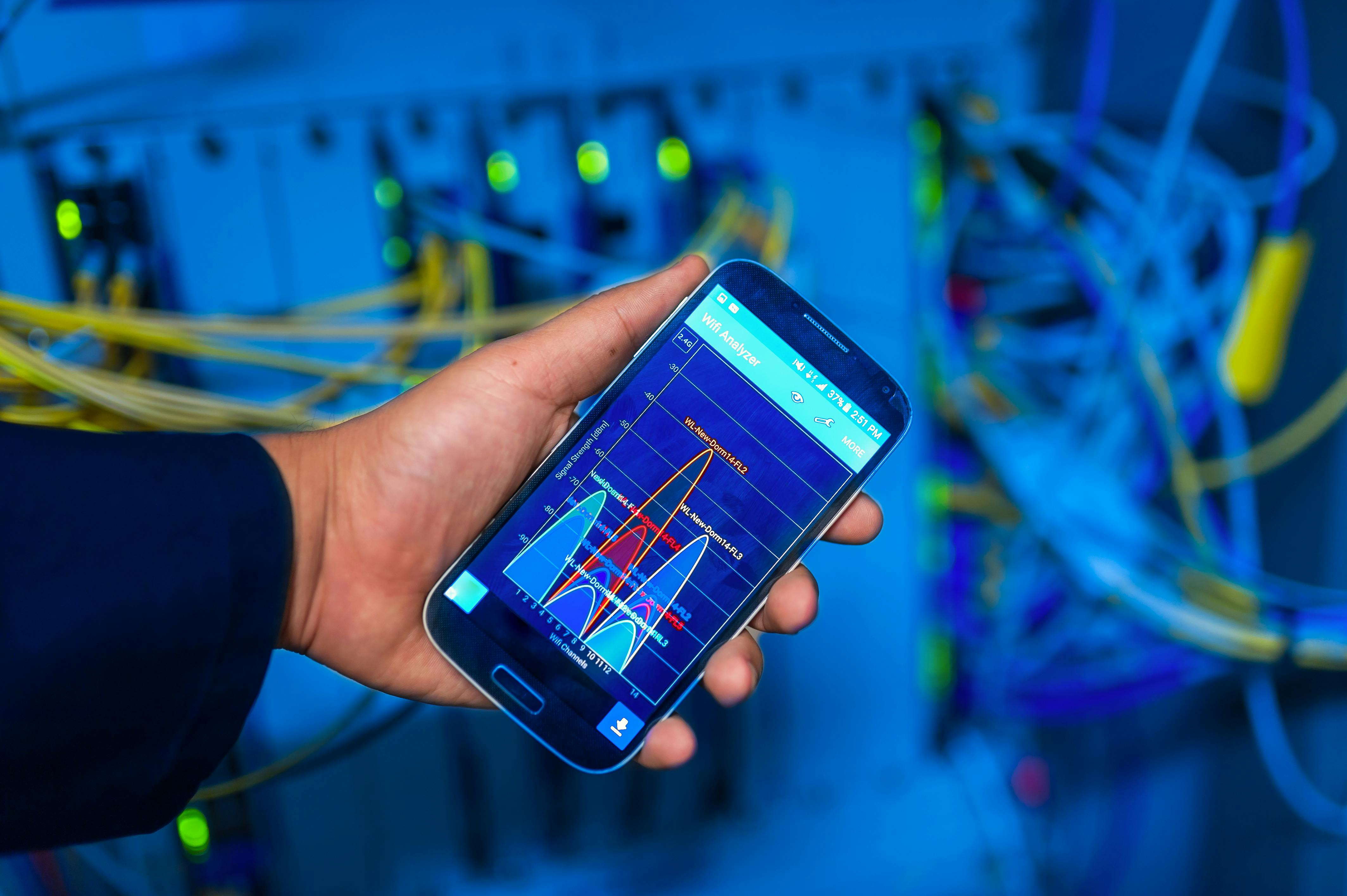 A person holds a smartphone displaying a graph, in front of network cables and equipment in a server room.