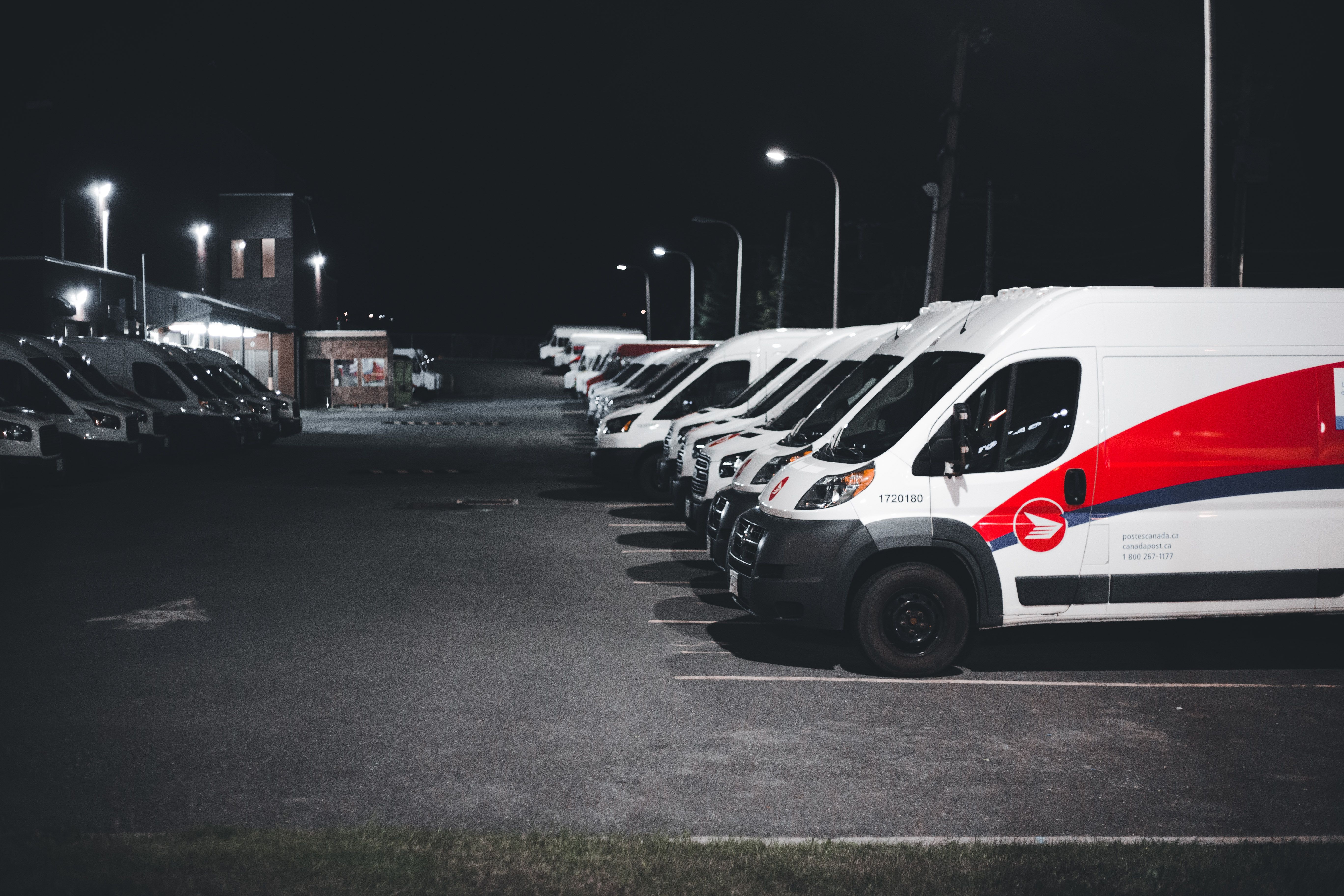 Row of postal delivery vans parked in a lot at night, with streetlights illuminating the area.