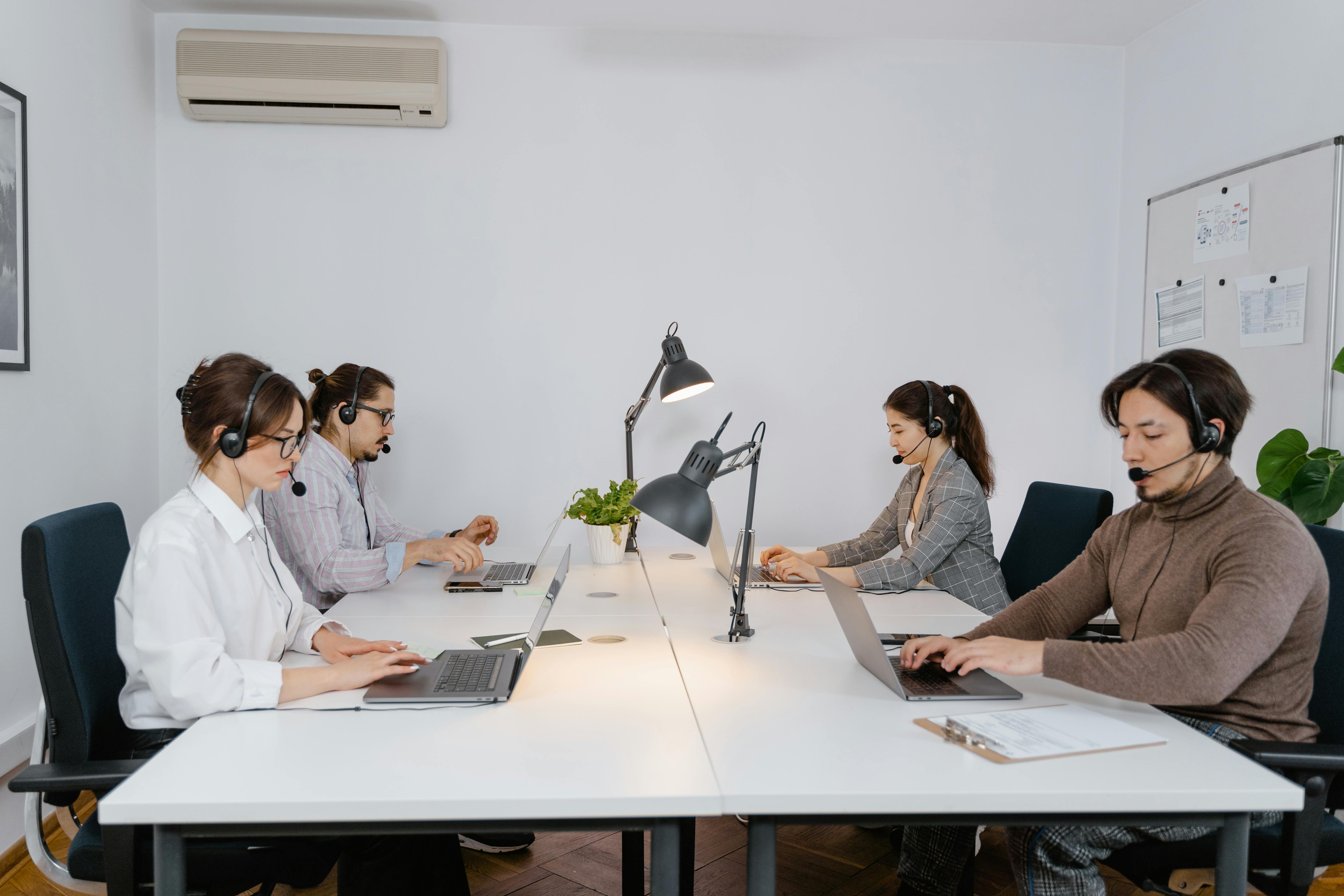 Four people wearing headsets work on laptops at a shared office table, with lamps and a small plant in the center.