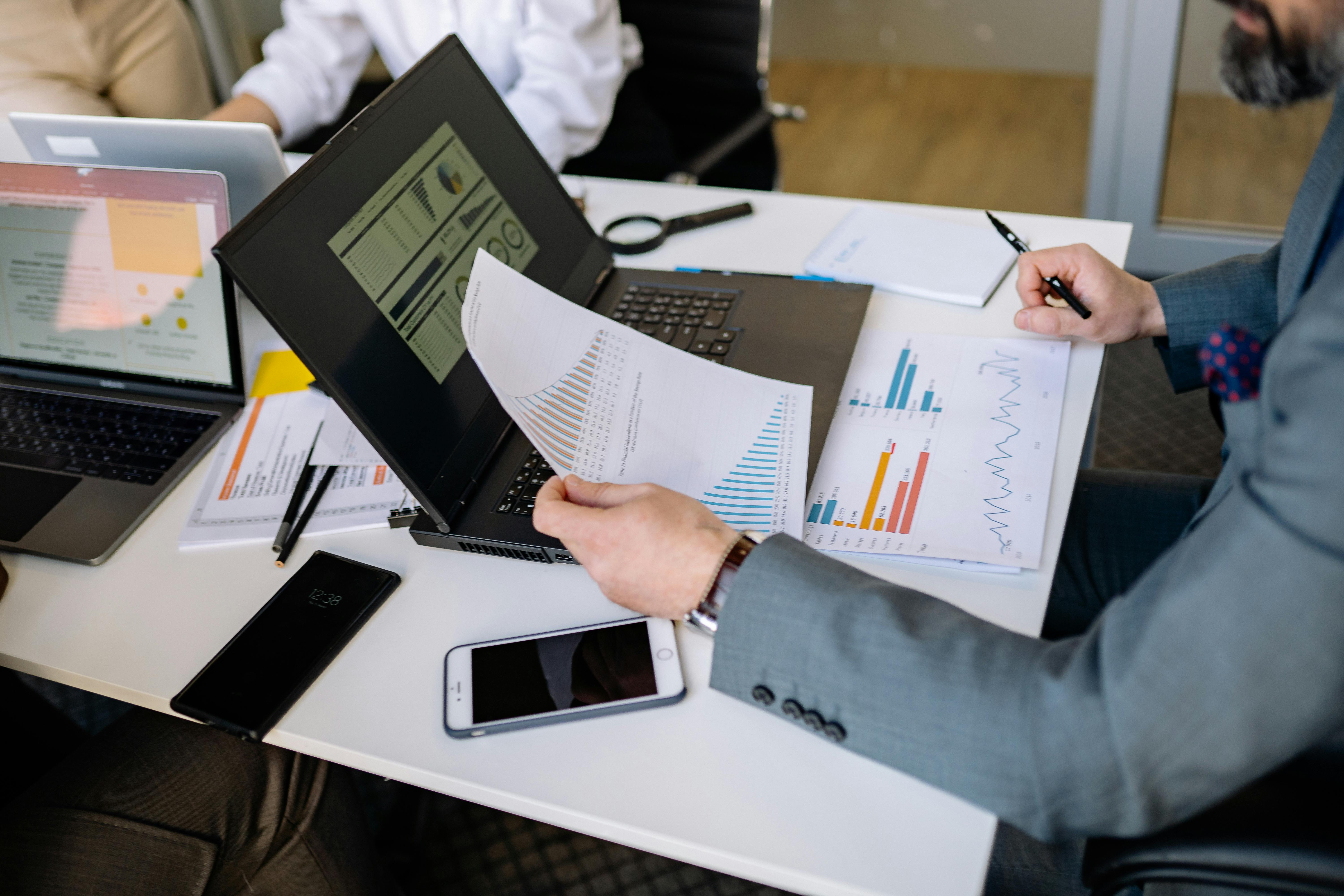 People at a desk analyzing charts and graphs on laptops and paper, with smartphones nearby, in a business meeting environment.