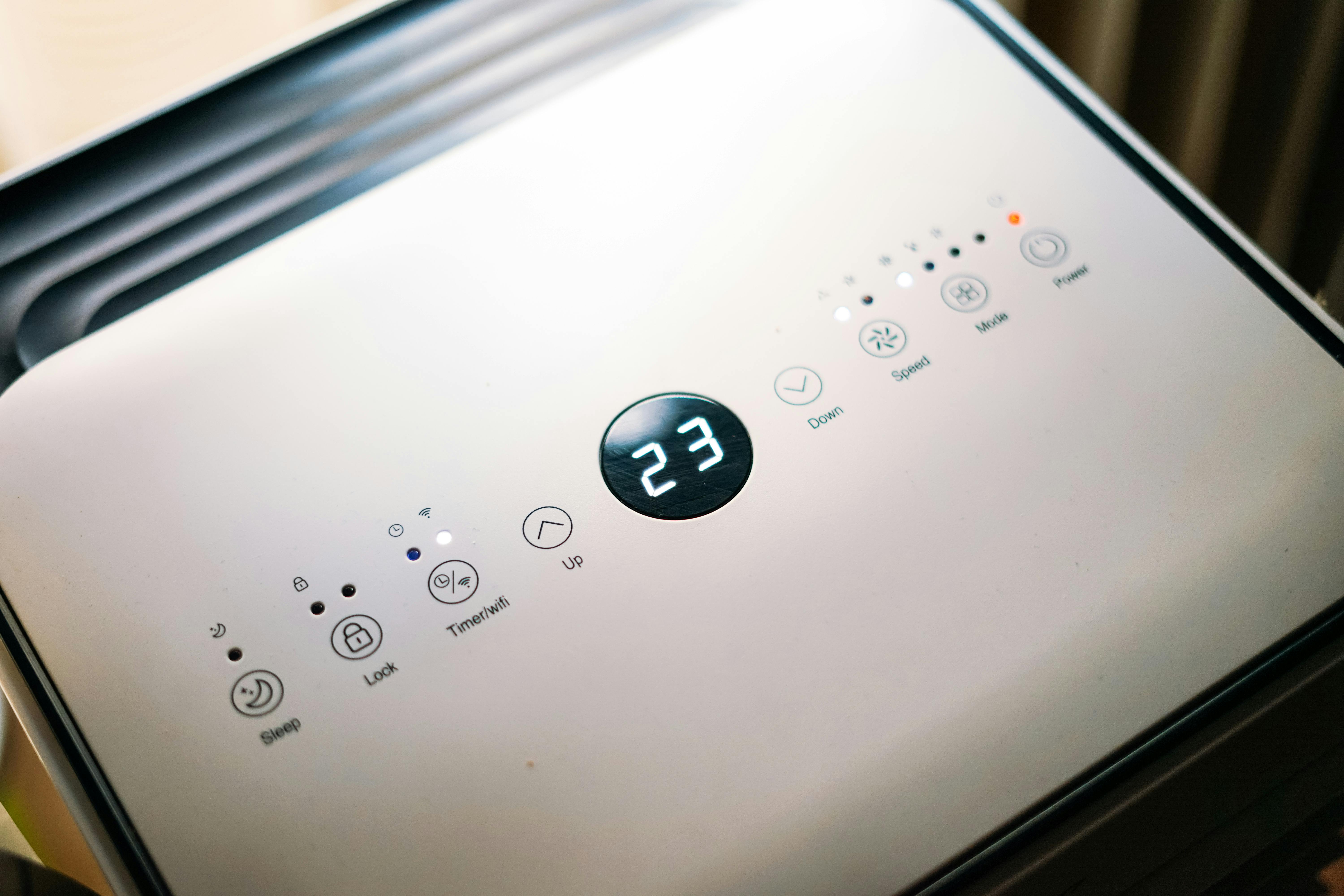 Close-up of an air purifier control panel displaying "23." Various function buttons and indicators are visible on the sleek white surface.