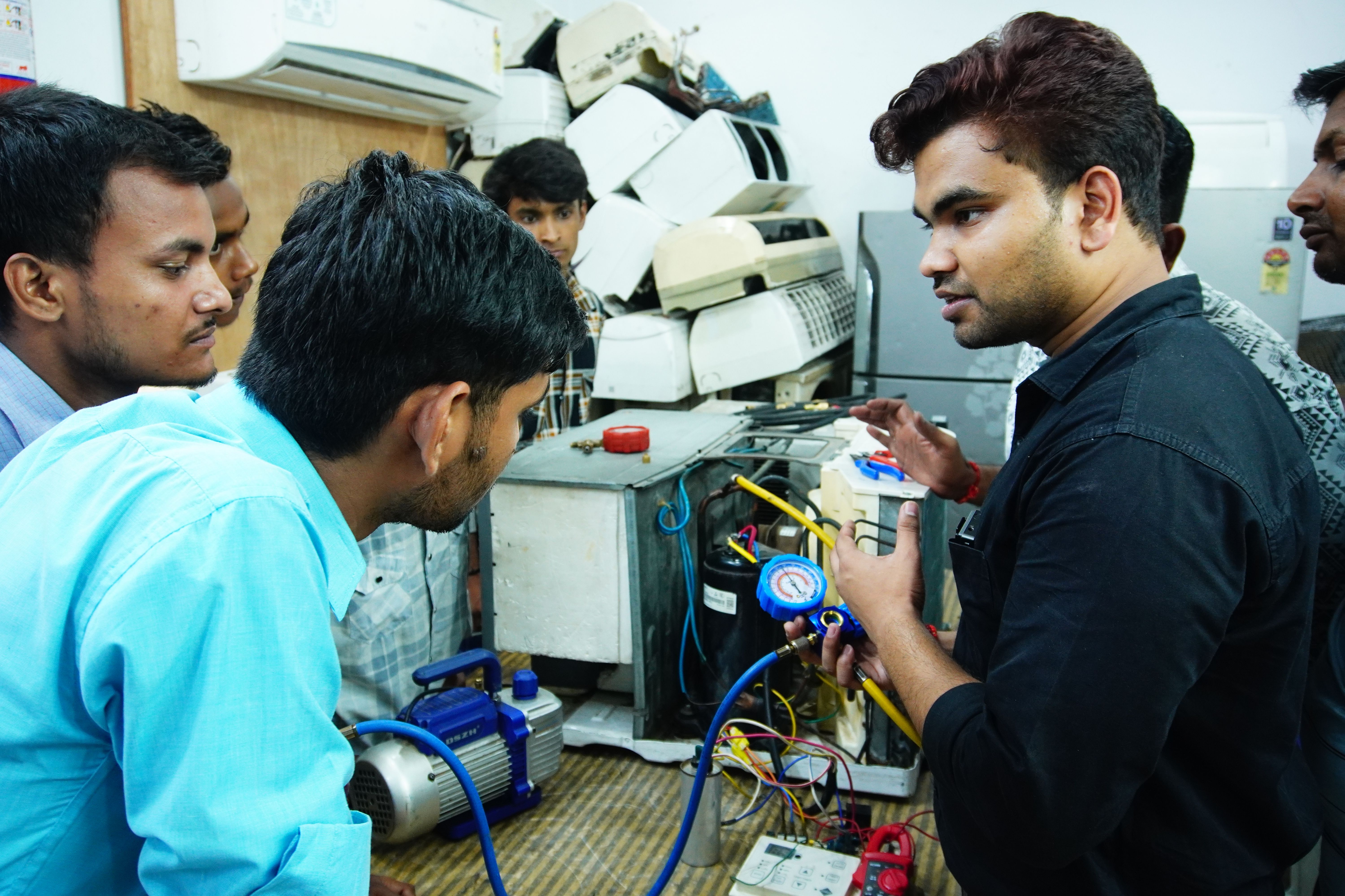 A group of men gathered around an instructor explaining a technical device, with various equipment visible in the background.