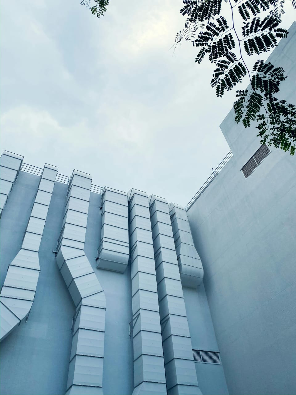 View of a building with vertical ducts against a cloudy sky, framed by green leaves.
