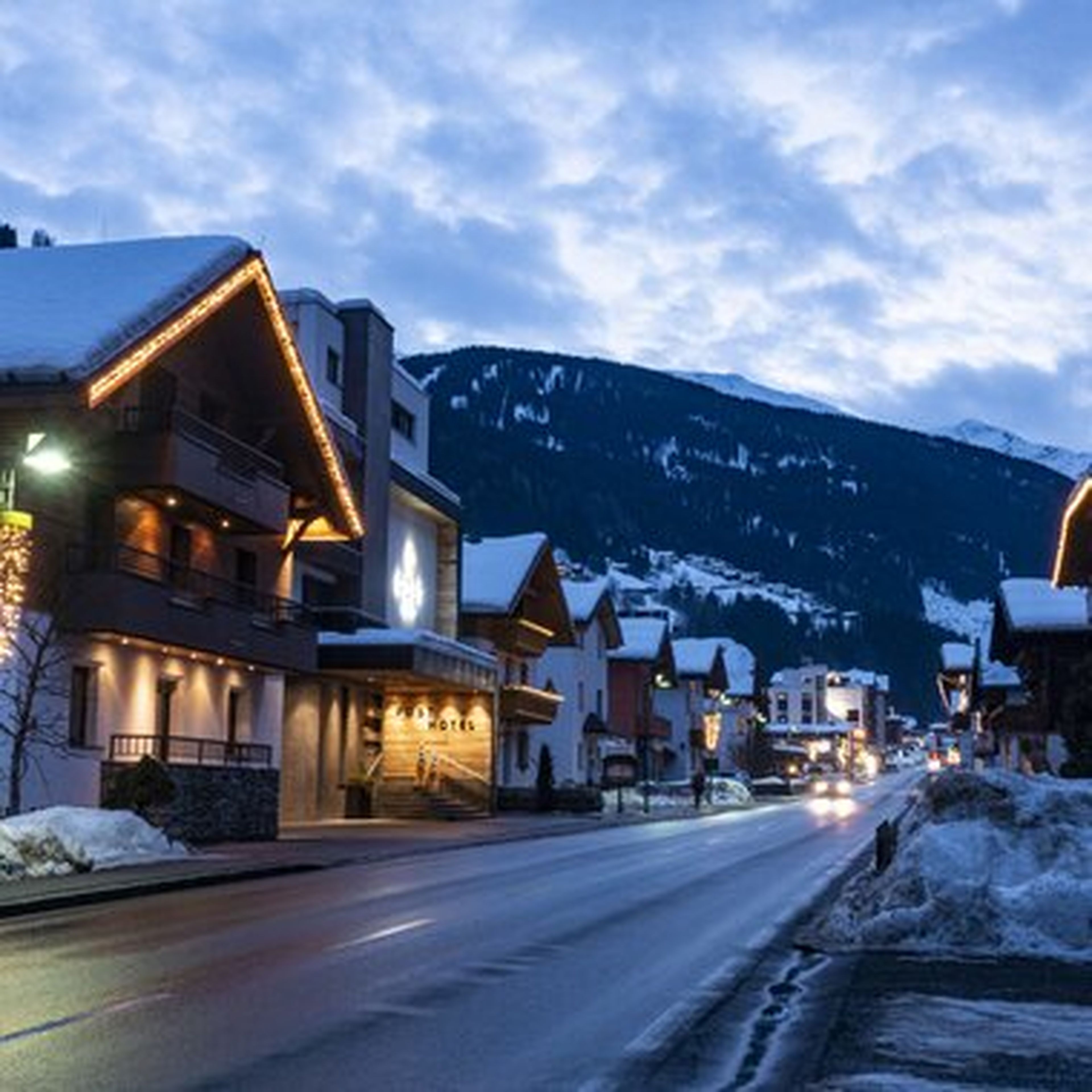 Verschneite Dorfstraße in den Alpen bei Dämmerung, mit einem hell beleuchteten Hotel, weiteren Gebäuden und dunklen Bergkonturen.