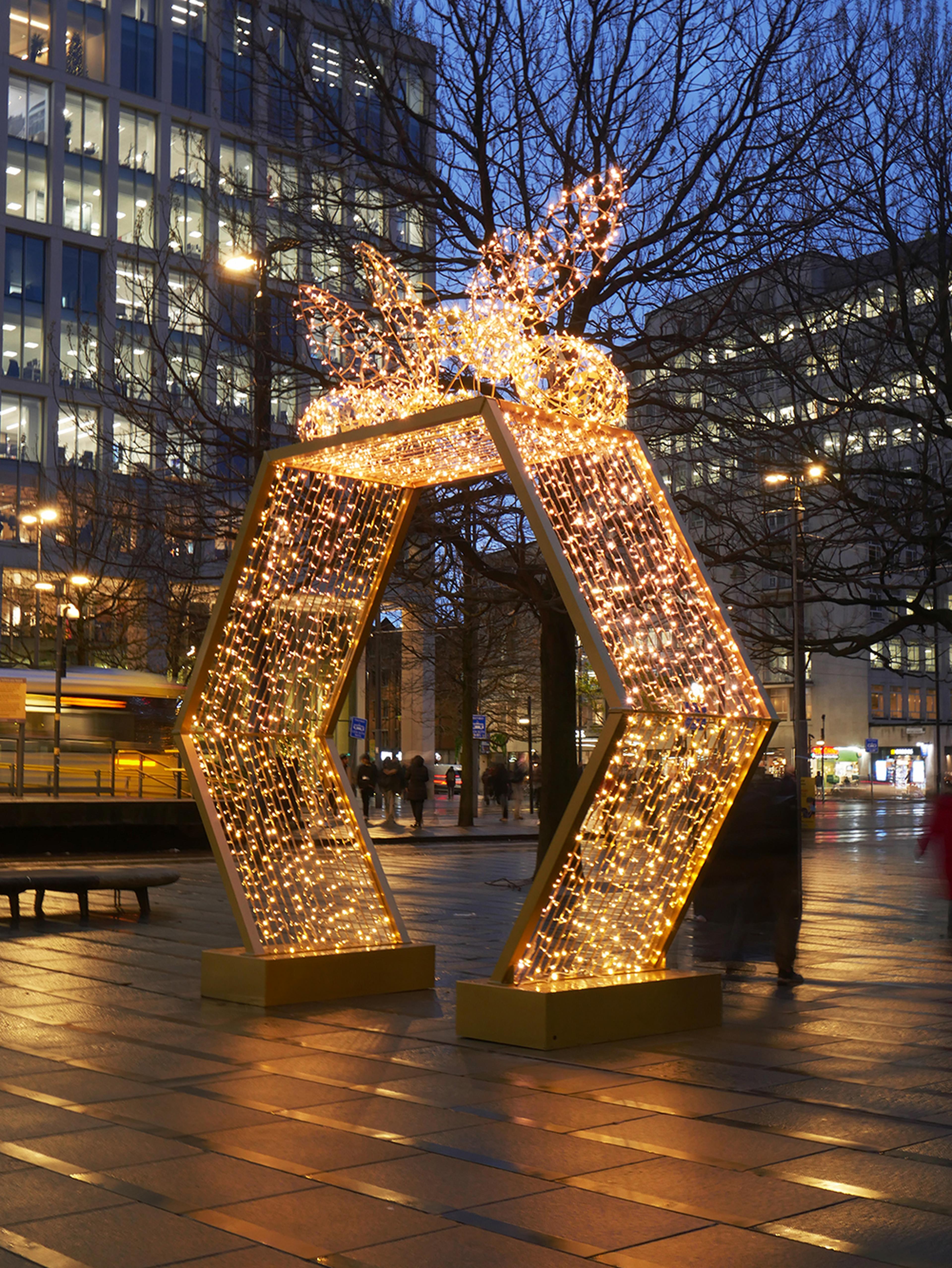 Leuchtende, sechseckige Lichtinstallation auf einem städtischen Platz bei Abenddämmerung, umgeben von hohen Gebäuden.
