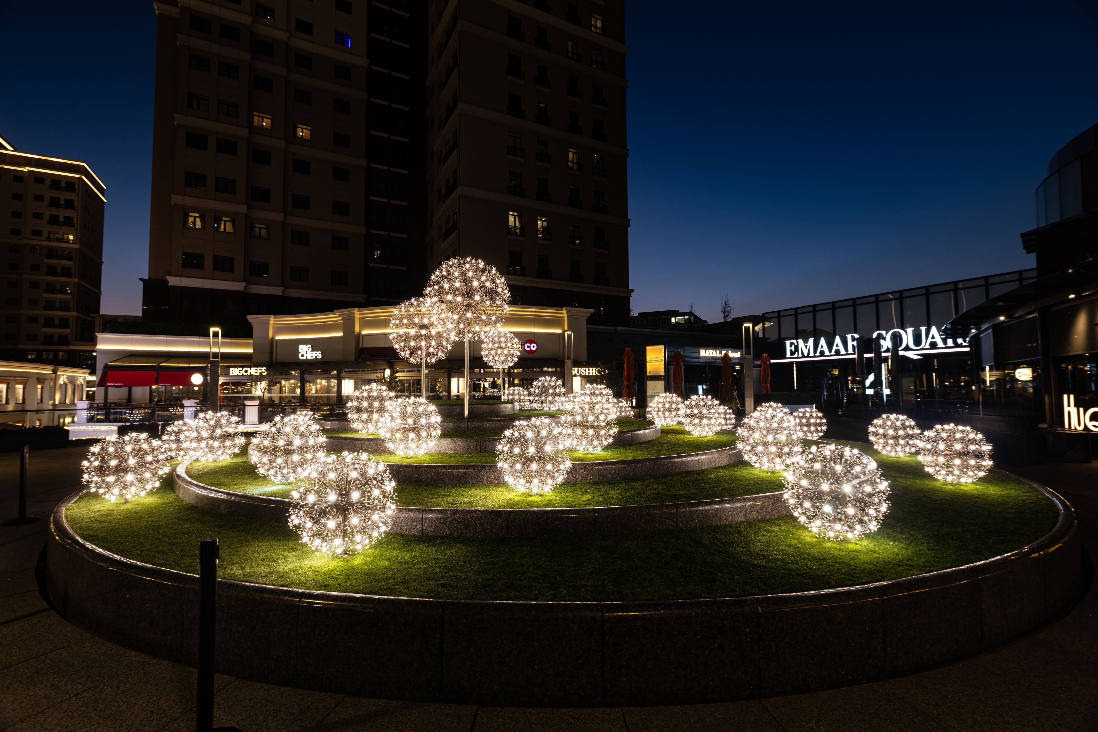 Kreisförmige Lichtskulpturen beleuchten eine abendliche, stufenförmig angelegte öffentliche Fläche, erzeugen ein festliches Ambiente.