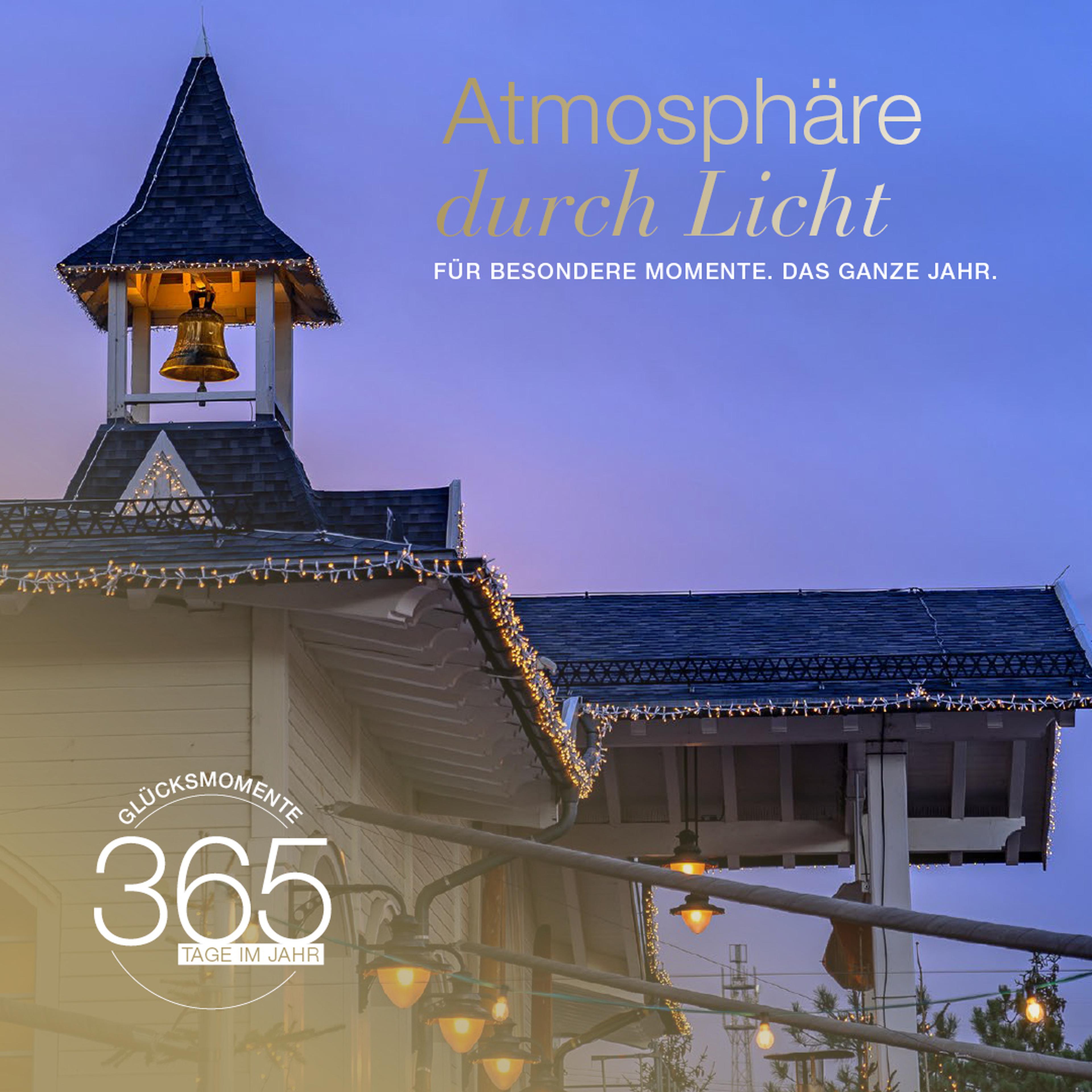 Dekoratives Gebäude mit Lichterketten, Glockenturm und Text "Atmosphäre durch Licht. Glücksmomente 365 Tage im Jahr" im Hintergrund.
