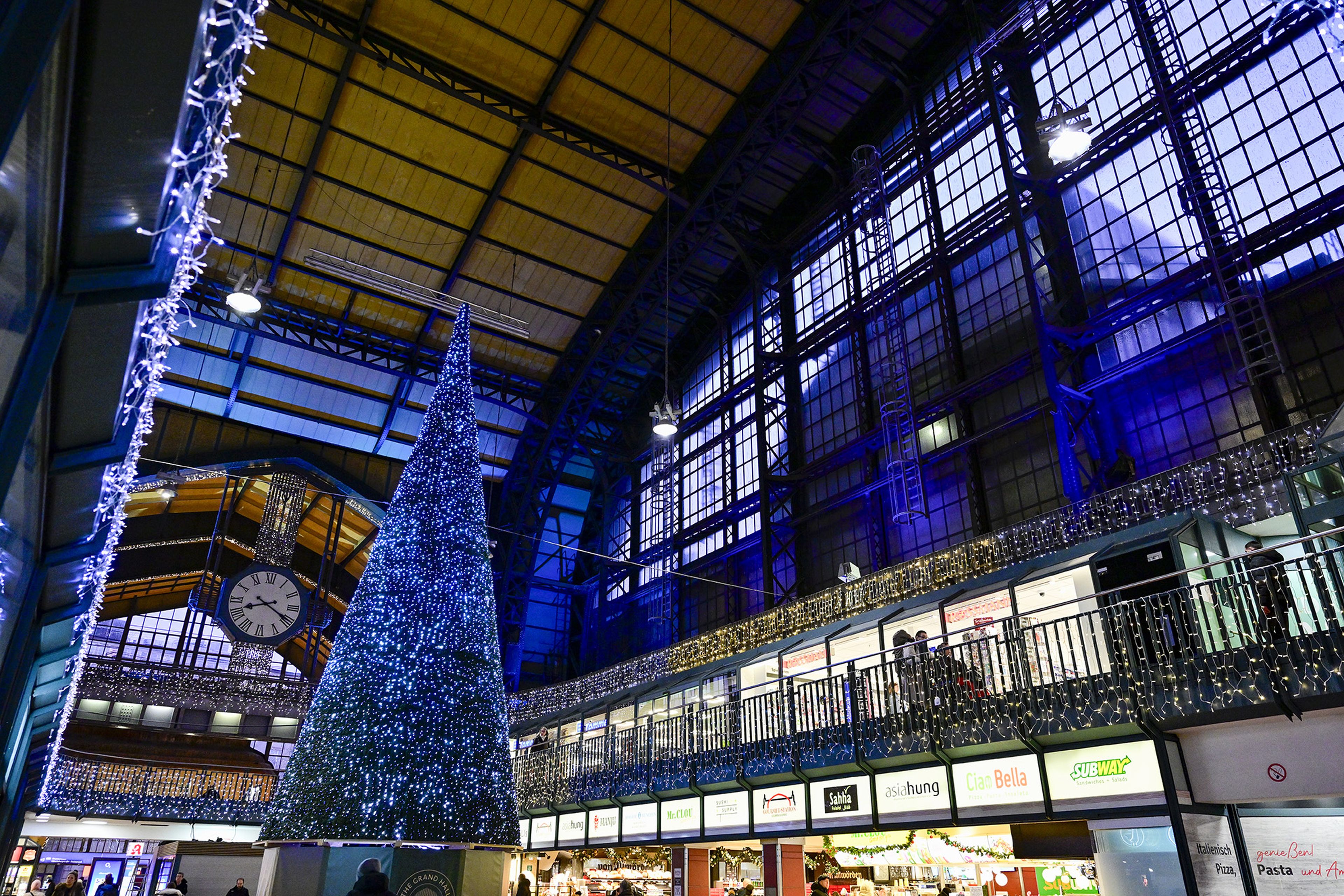 Festlich erleuchteter Bahnhof mit hohem Weihnachtsbaum und Glühlampenketten an Geländern; Geschäfte im Hintergrund.