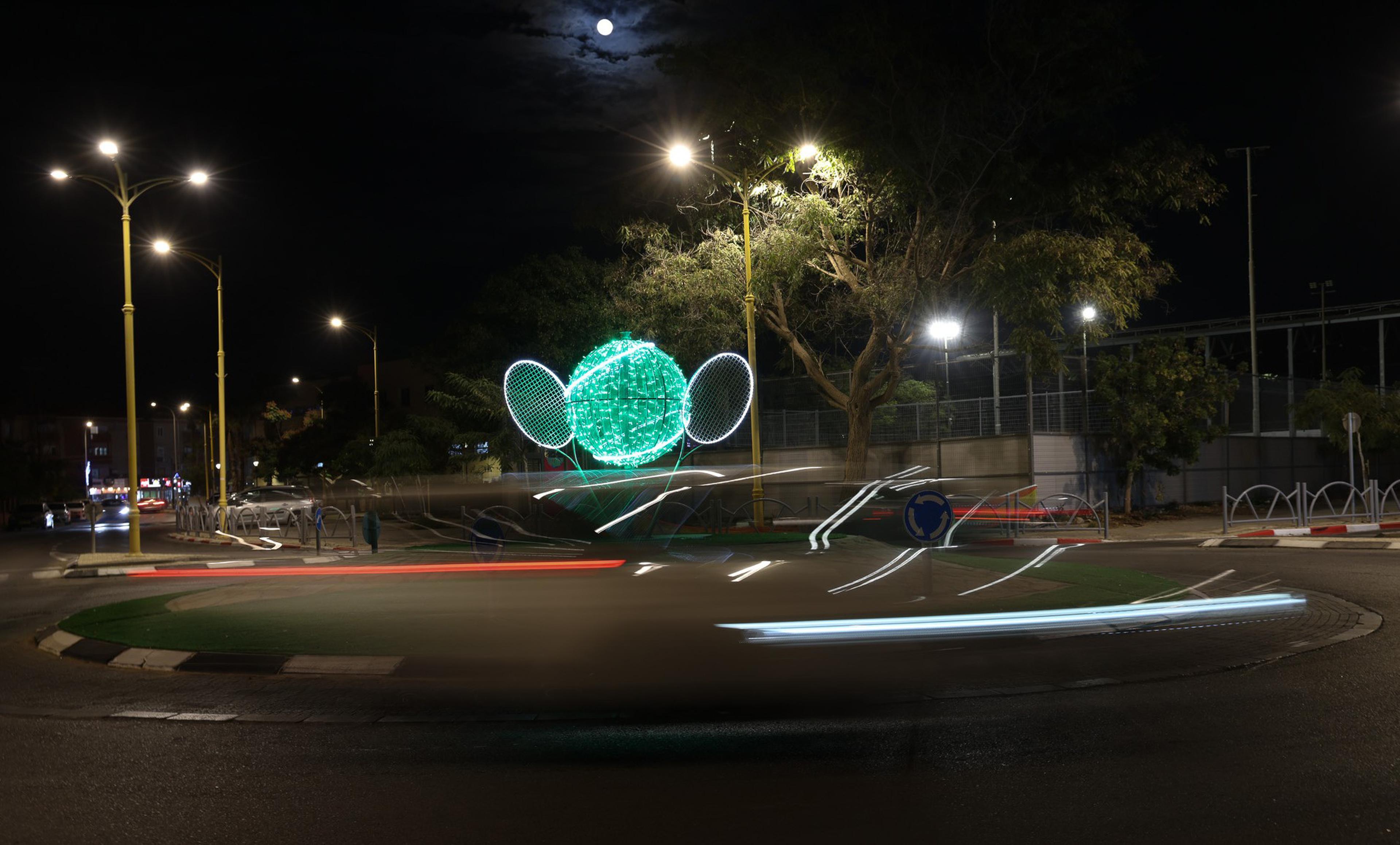 Runde Straßenkunst aus grünem Licht mit Tennismotiven in einem Kreisverkehr bei Nacht, umgeben von Straßenlichtern.