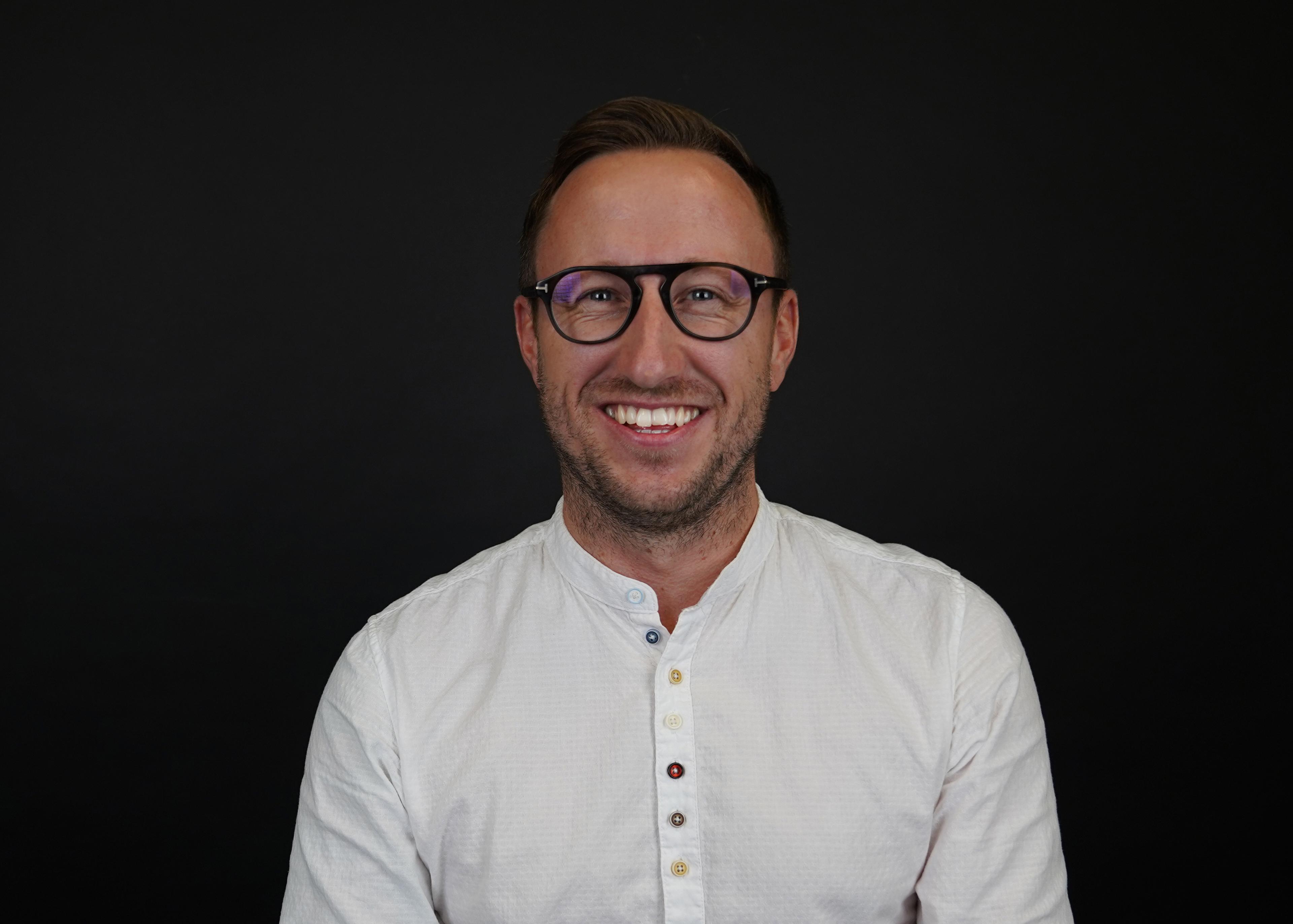 Lächelnder Mann mit Brille und weißem Hemd vor schwarzem Hintergrund.