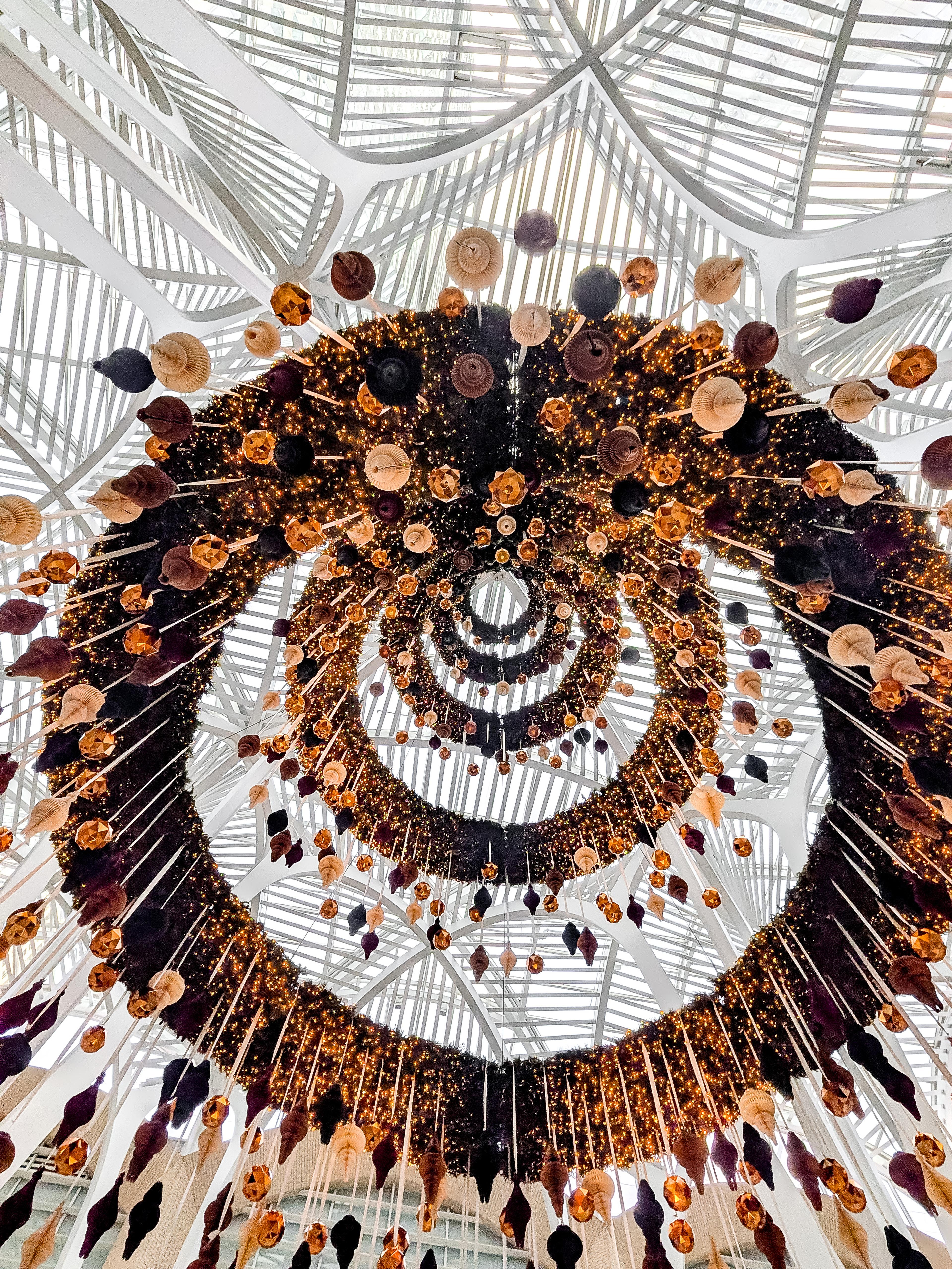 Aufwendige Deckeninstallation mit kreisförmigen Ornamenten und Lichtern in warmen Tönen, vor einer modernen, gewölbten Glasdecke.