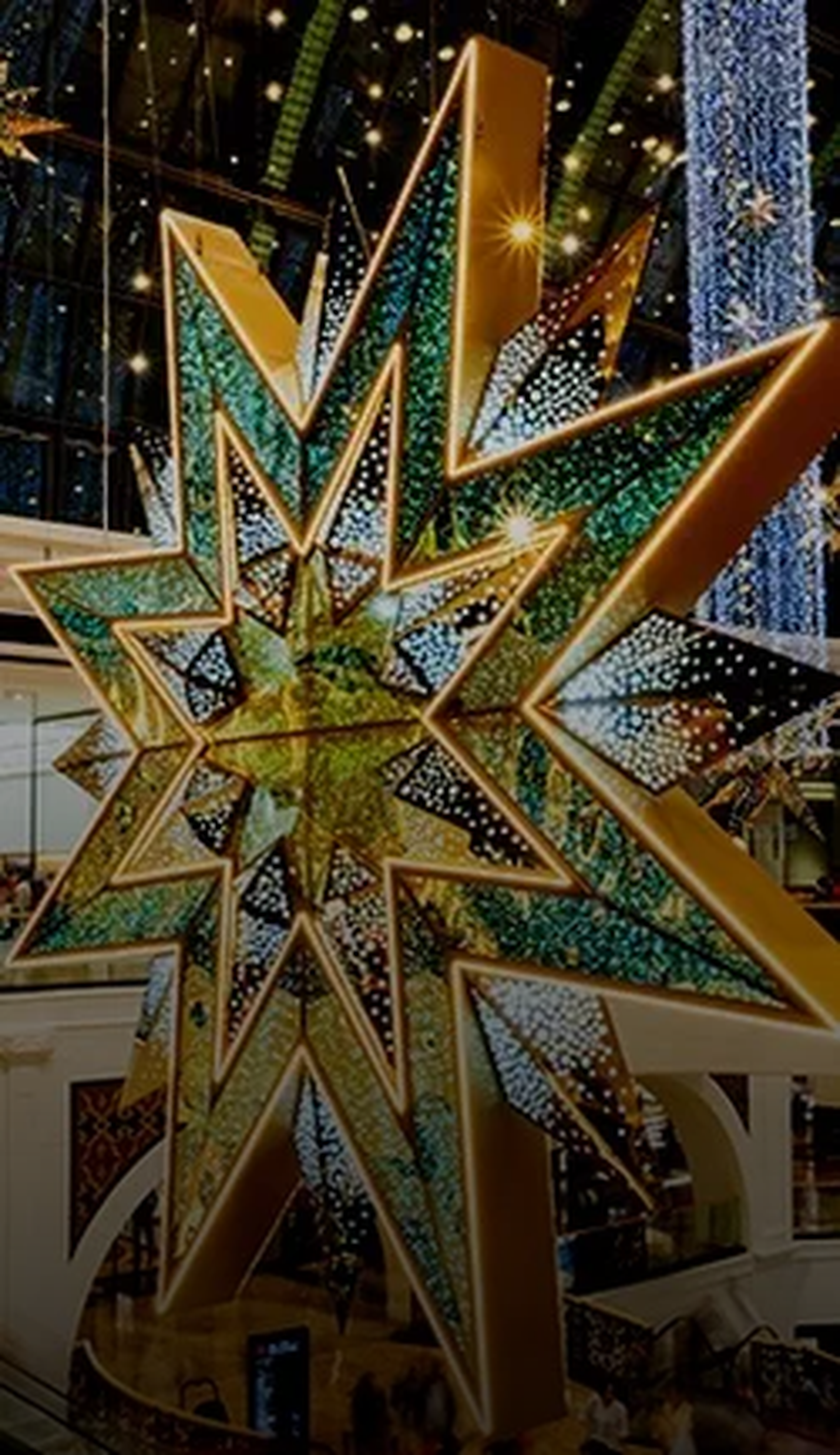 Großer, leuchtender Weihnachtsstern aus goldenen und grünen Elementen, hängt in einem Einkaufszentrum, umgeben von weiteren dekorativen Lichtern.