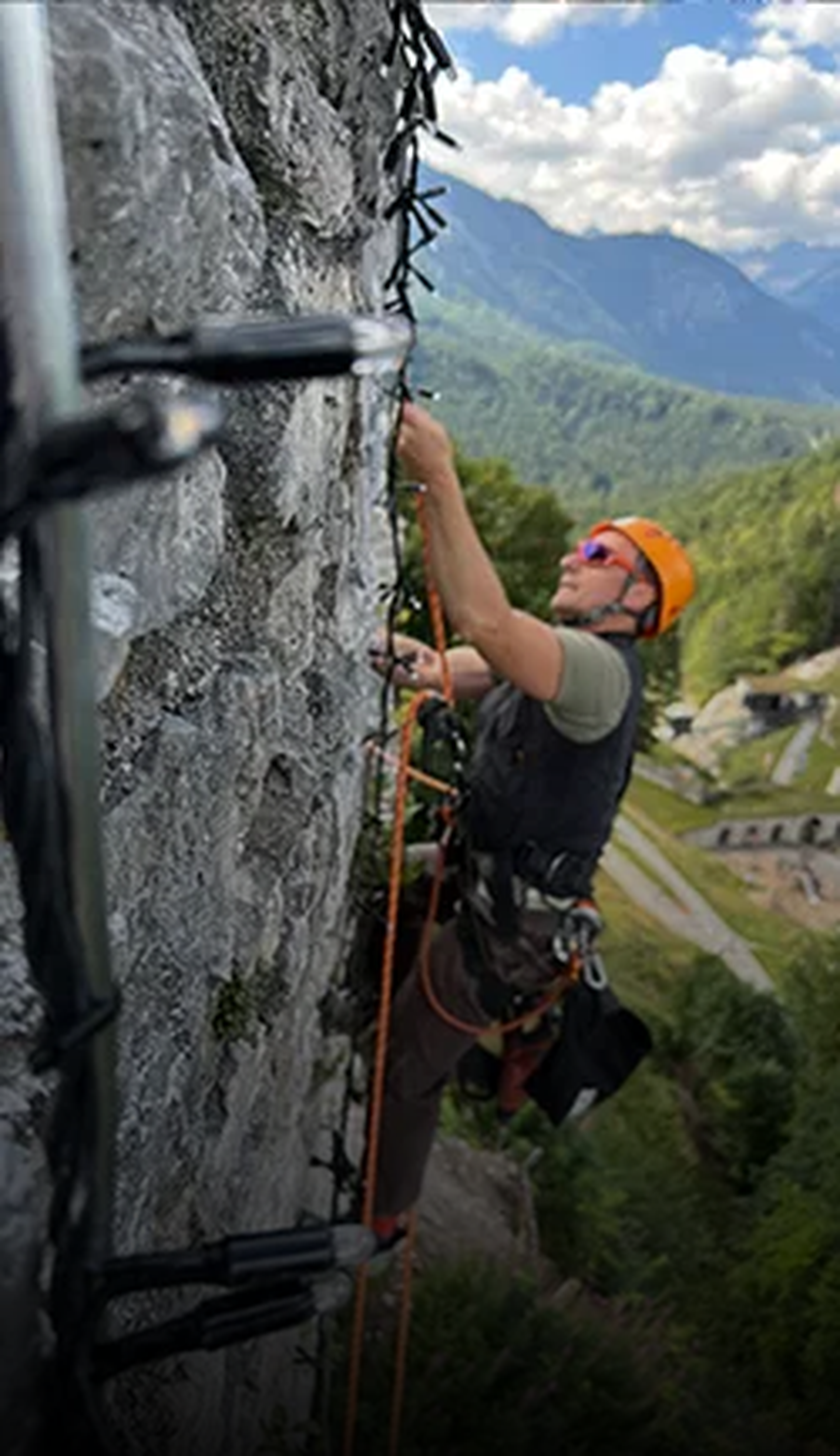 Ein Arbeiter mit Helm klettert eine Felswand hoch, befestigt Lichterketten; Berglandschaft im Hintergrund.