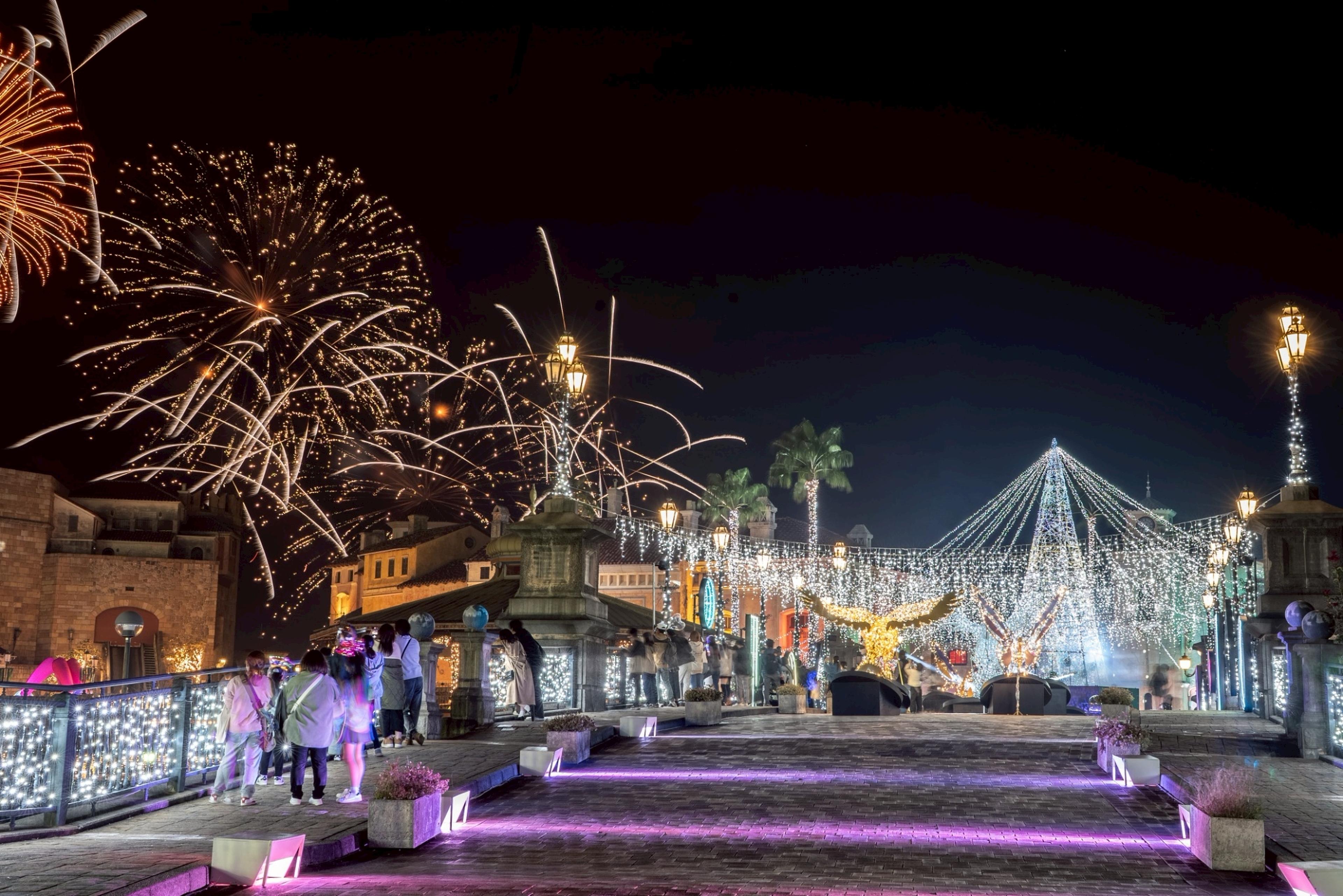 Festlich beleuchtete Straße mit Lichterketten, Menschen und Feuerwerk am Nachthimmel, Palmen und historisch anmutende Gebäude im Hintergrund.