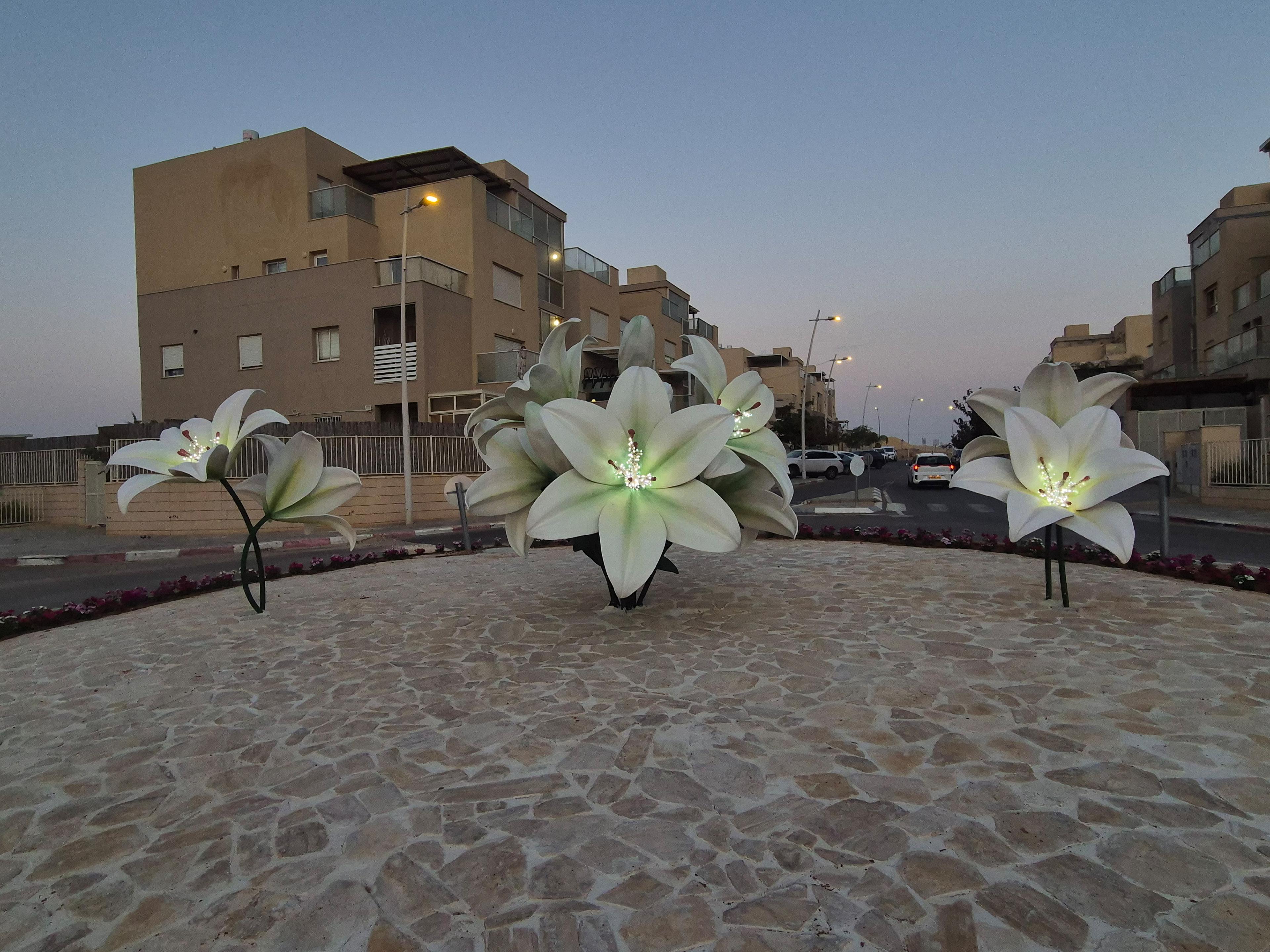 Große, beleuchtete Lilienfiguren in einem Kreisverkehr bei Dämmerung, umgeben von Wohngebäuden und einer gepflasterten Oberfläche.