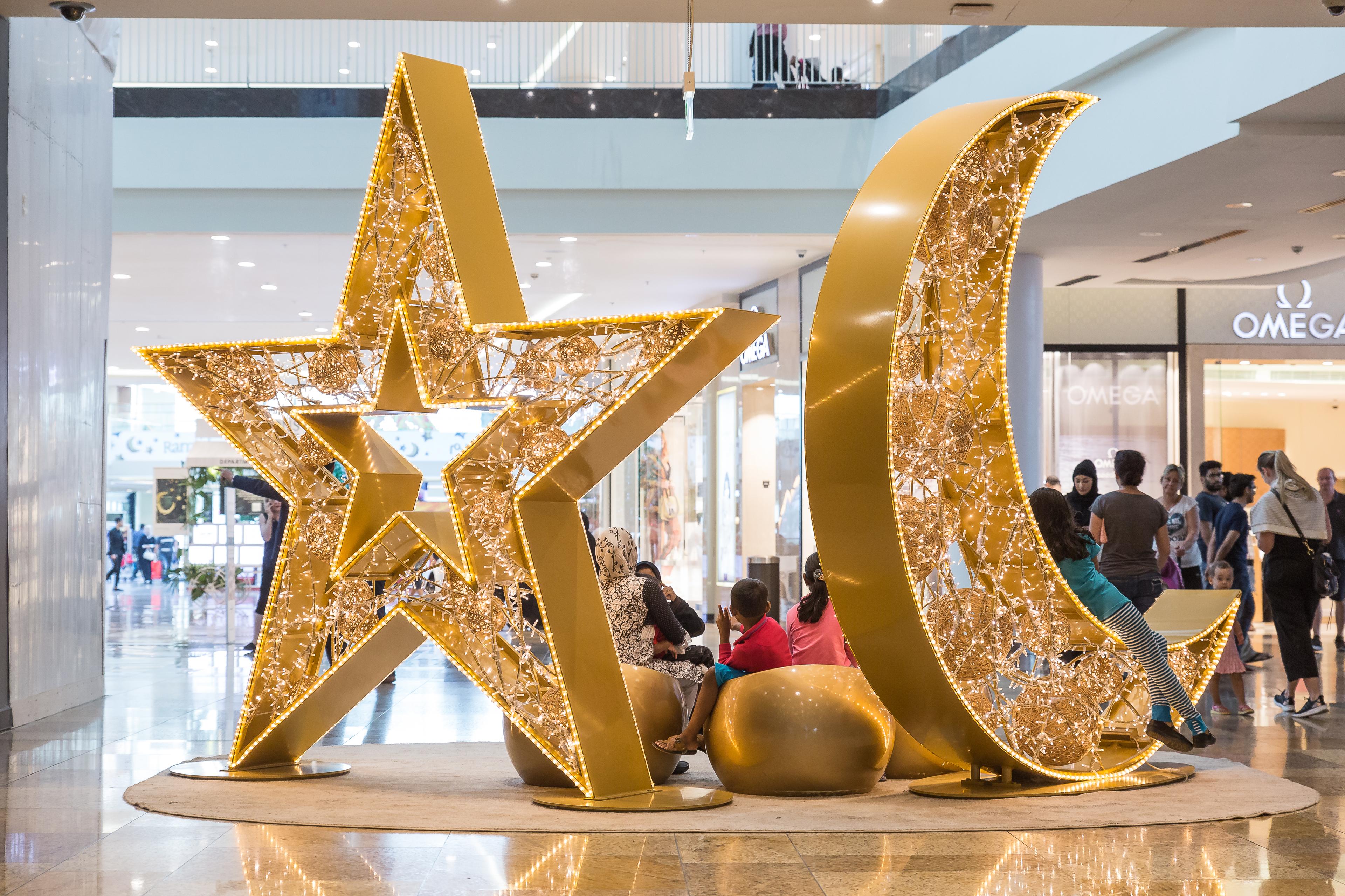 Goldene Stern- und Mondsitzgelegenheiten mit LED-Lichtern in einem Einkaufszentrum, umgeben von Menschen.