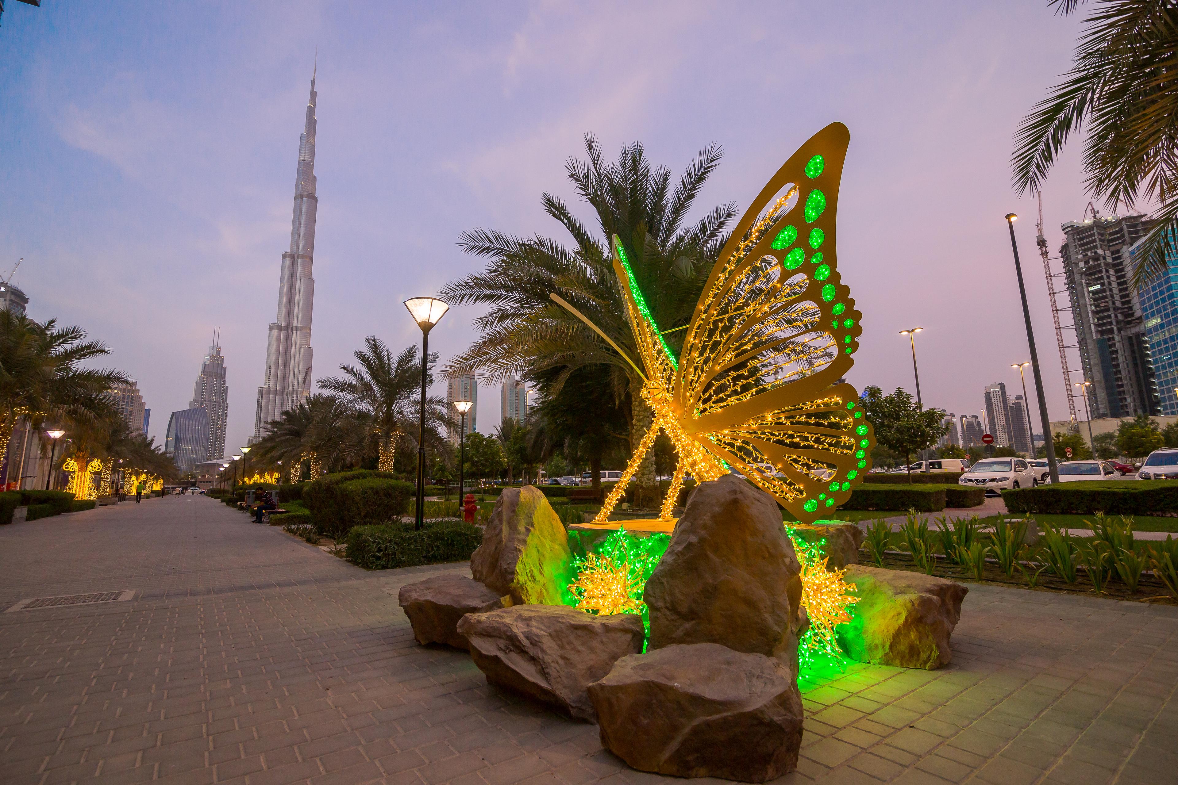 Glitzernde Schmetterlingsskulptur mit Beleuchtung in einem urbanen Park bei Abenddämmerung, im Hintergrund ein hoher Turm.