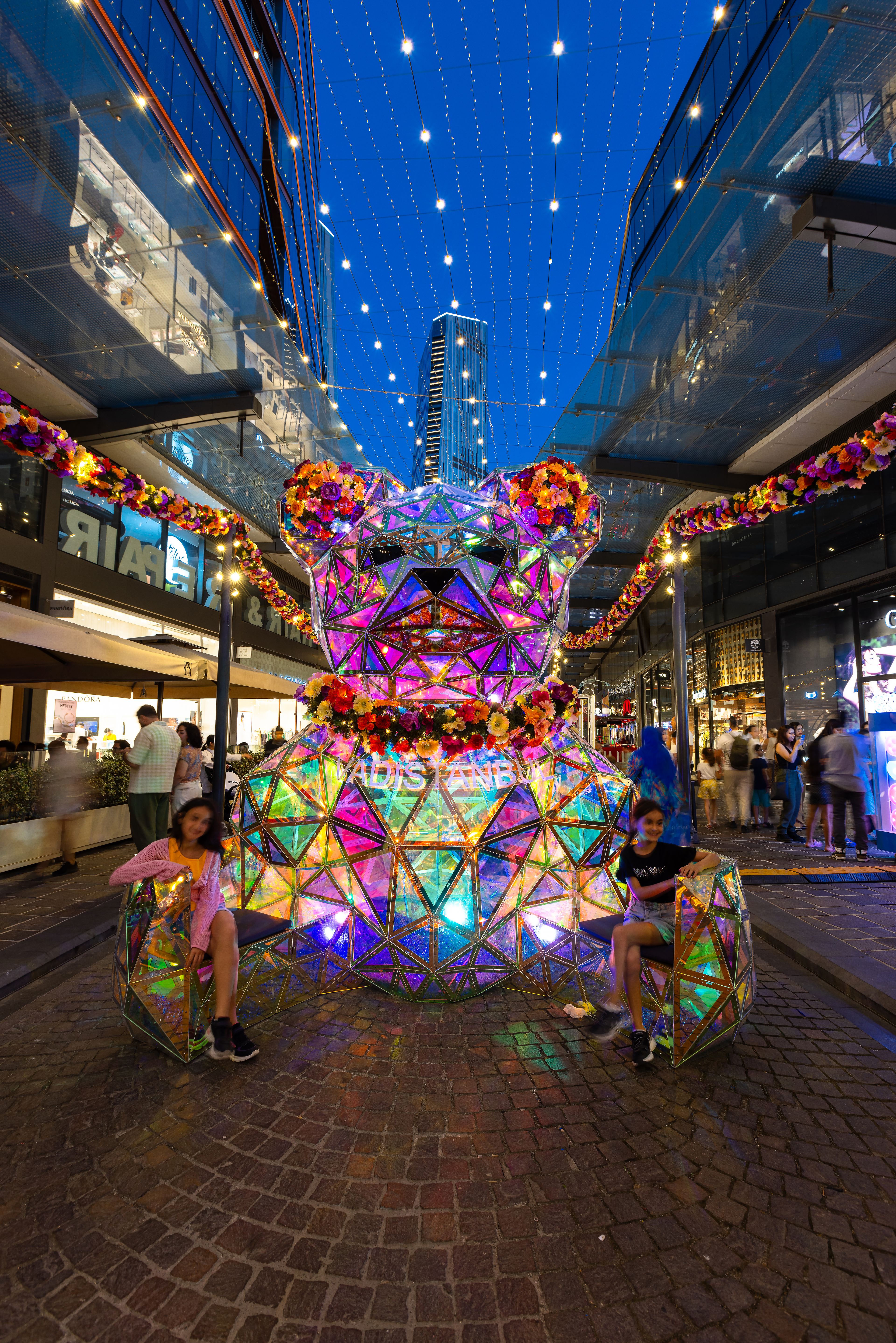 Lichterbär mit bunter Blumenpracht beleuchtet städtischen Platz bei Nacht, umgeben von Passanten.