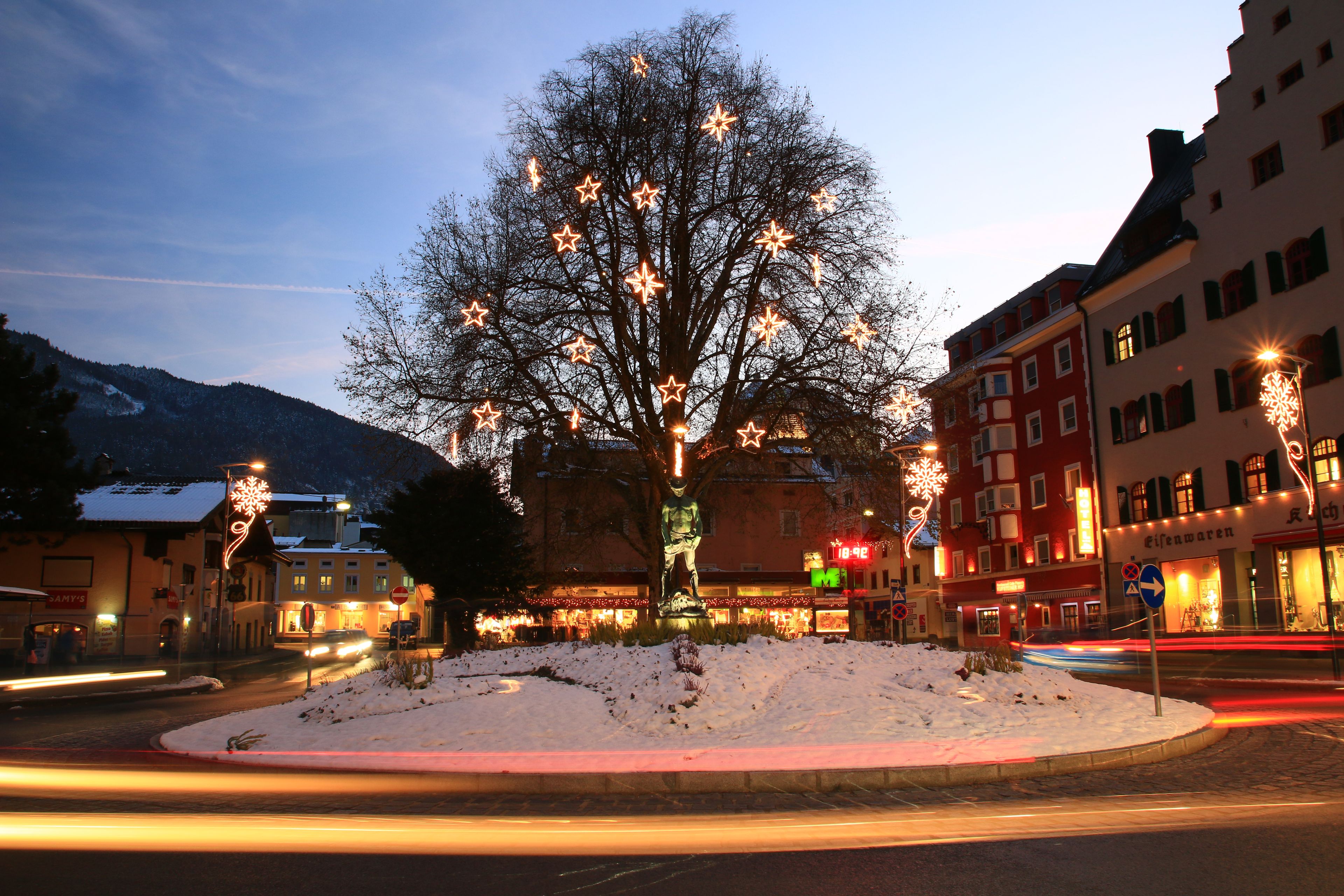 Beleuchteter Baum auf einem schneebedeckten Kreisverkehr bei Abenddämmerung, umgeben von Gebäuden und leuchtenden Sternen.