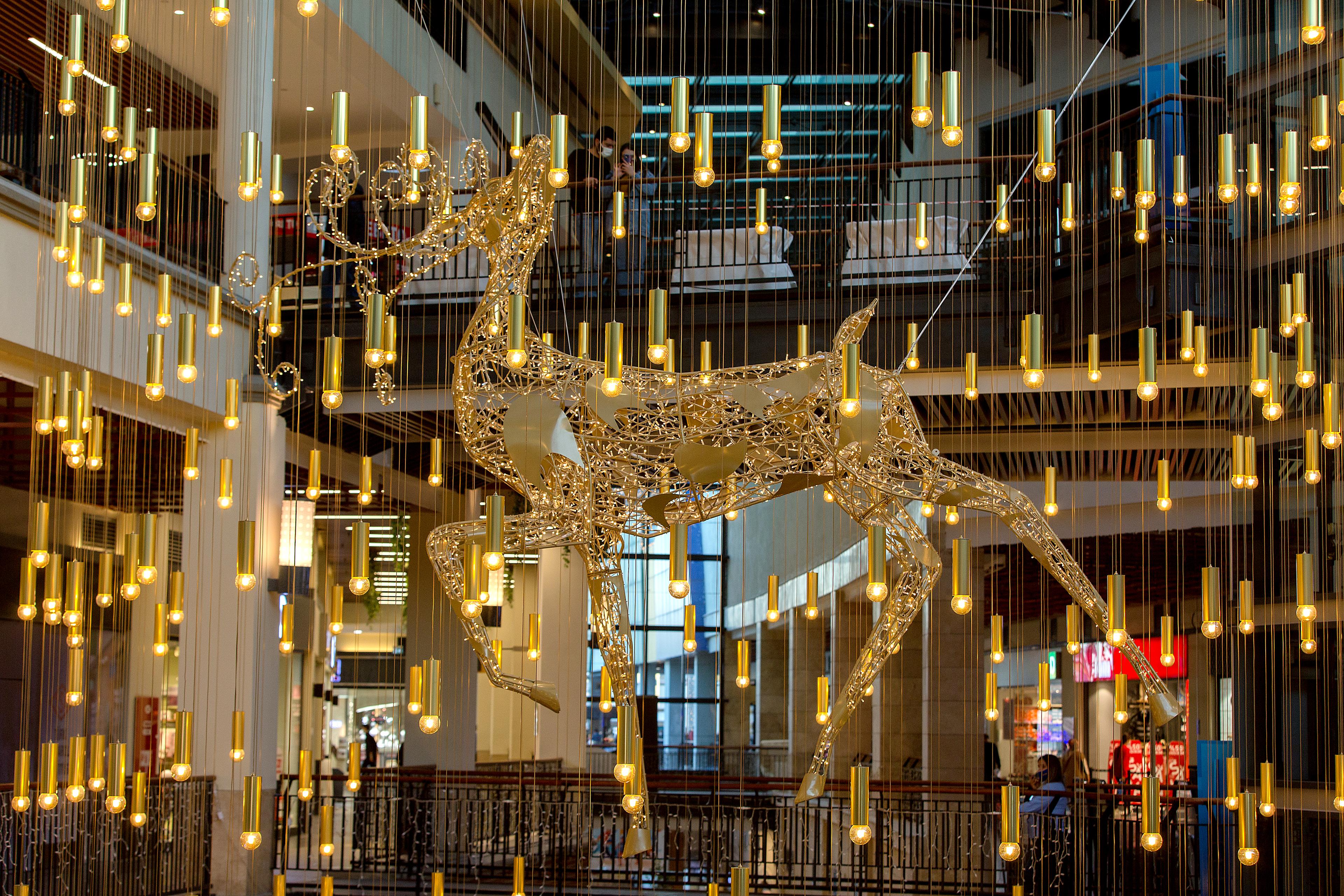 Goldfarbener Rentier-Leuchtskulptur hängt in einem Einkaufszentrum, umgeben von zahlreichen hängenden Lichtdekorationen.
