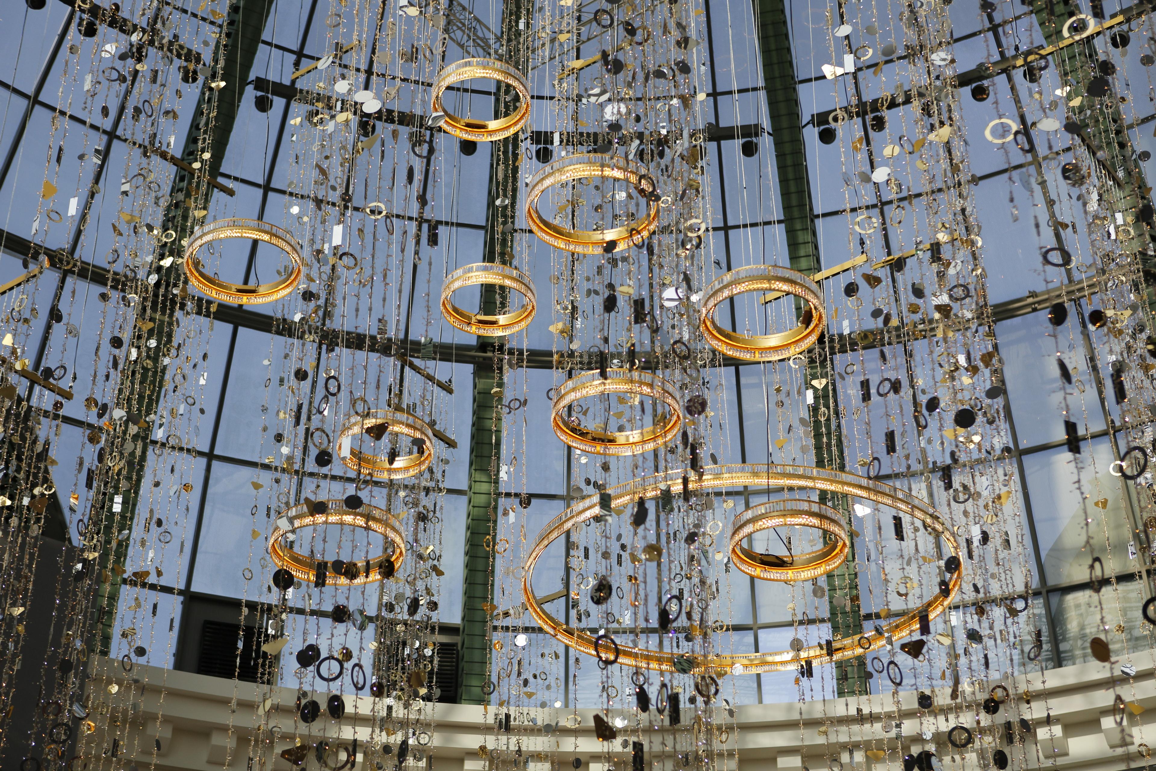 Kunstvolle hängende Lichtinstallation mit goldenen Ringen und funkelnden Dekorationen in einem gläsernen Gebäude.
