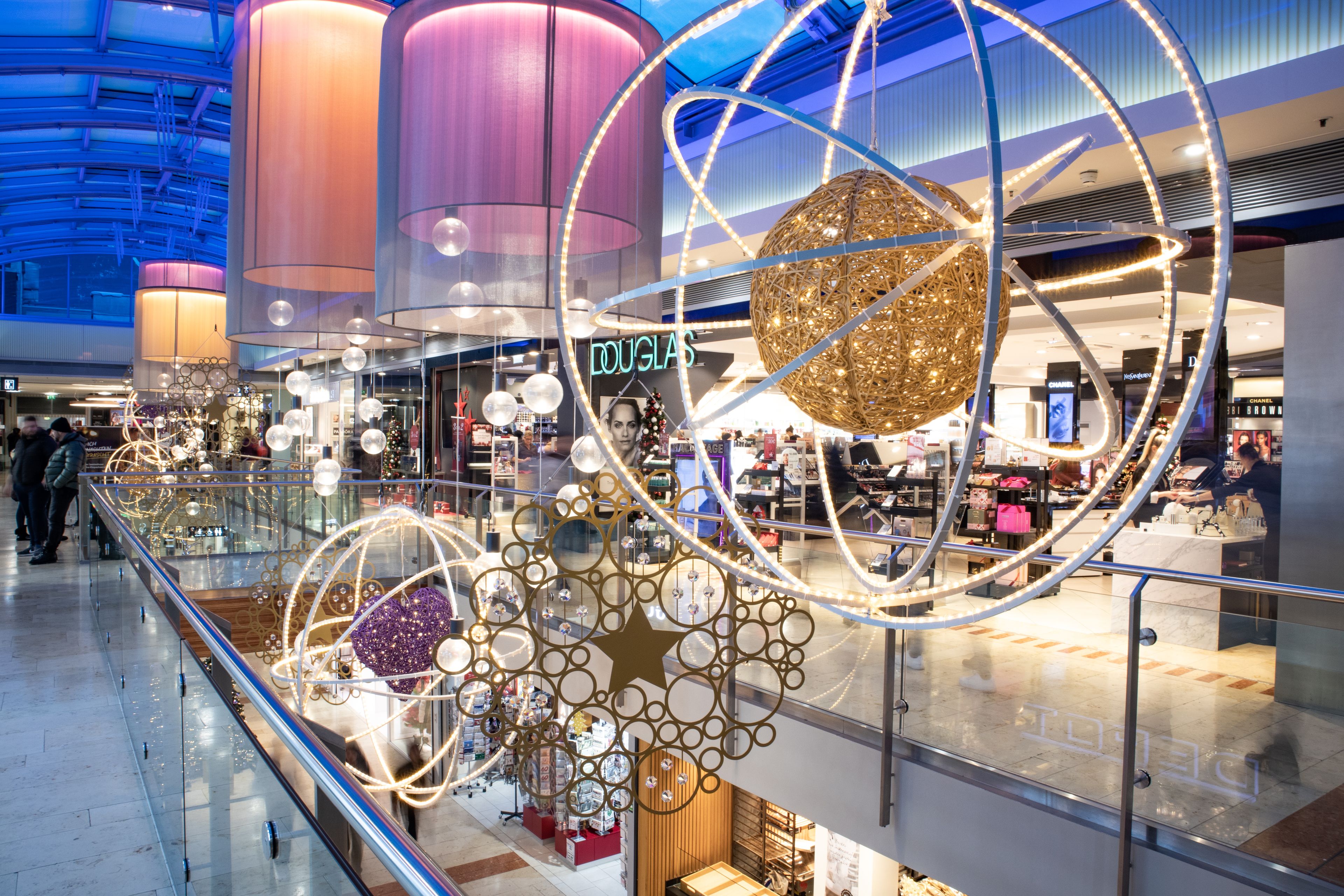 Einkaufszentrum mit festlicher Beleuchtung, großen leuchtenden Kugeln und modernen Dekorationen an einem Geländer im Innenbereich.