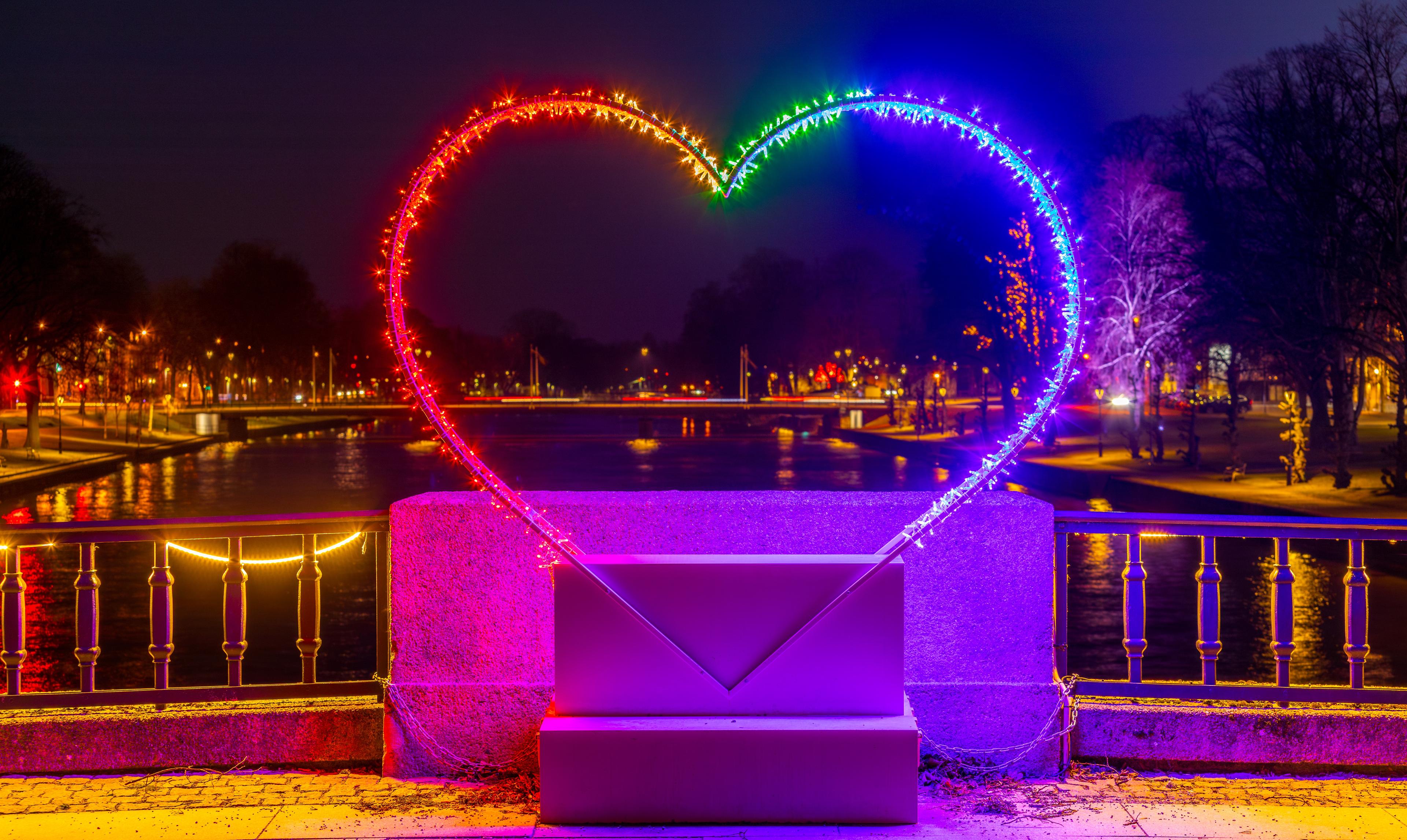 Beleuchtetes Herz in Regenbogenfarben auf Brücke bei Nacht, mit reflektierendem Wasser im Hintergrund.