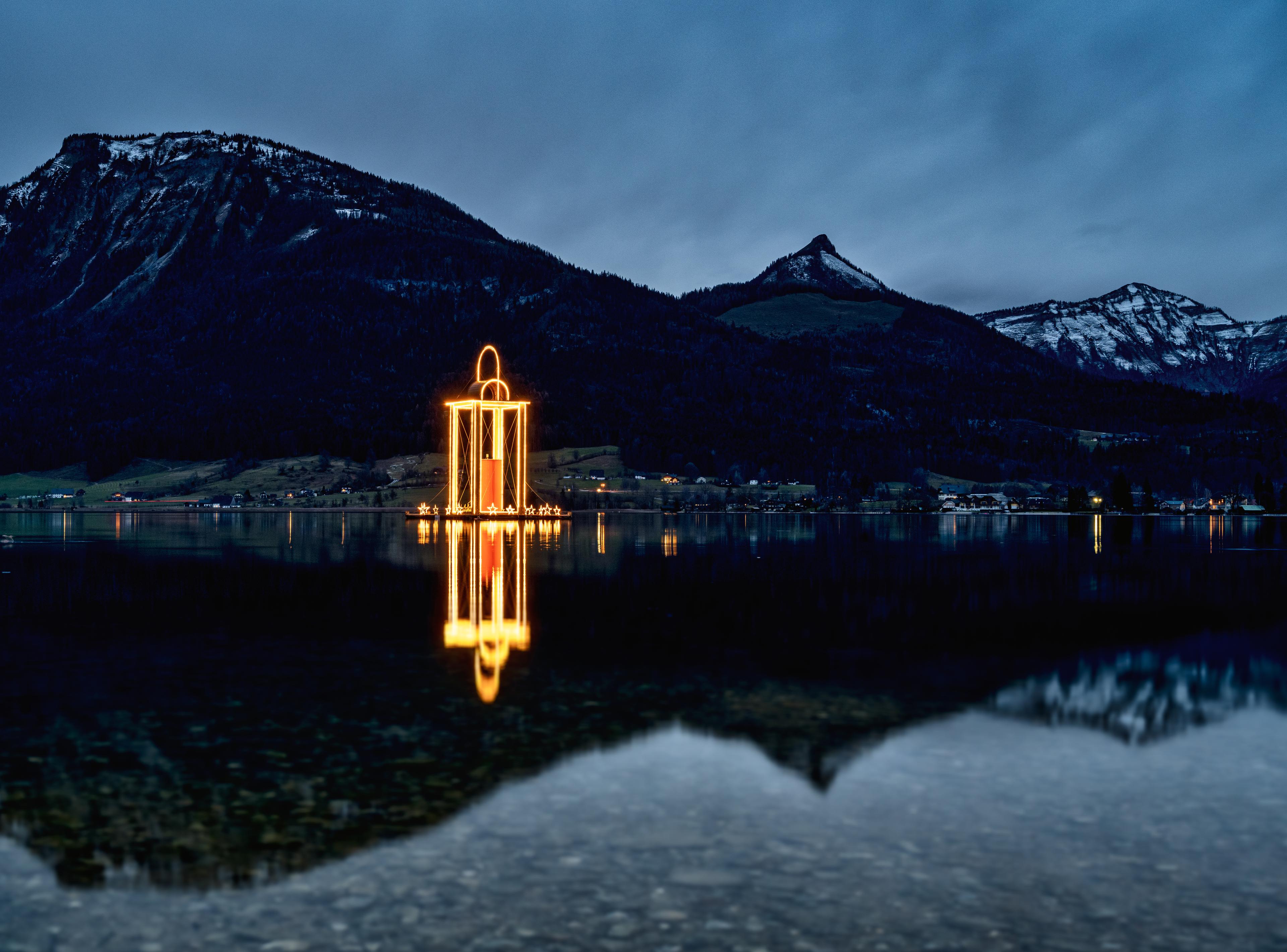 Leuchtendes Laternenkunstwerk spiegelt sich auf einem ruhigen See, umgeben von dunklen Bergen im Hintergrund.
