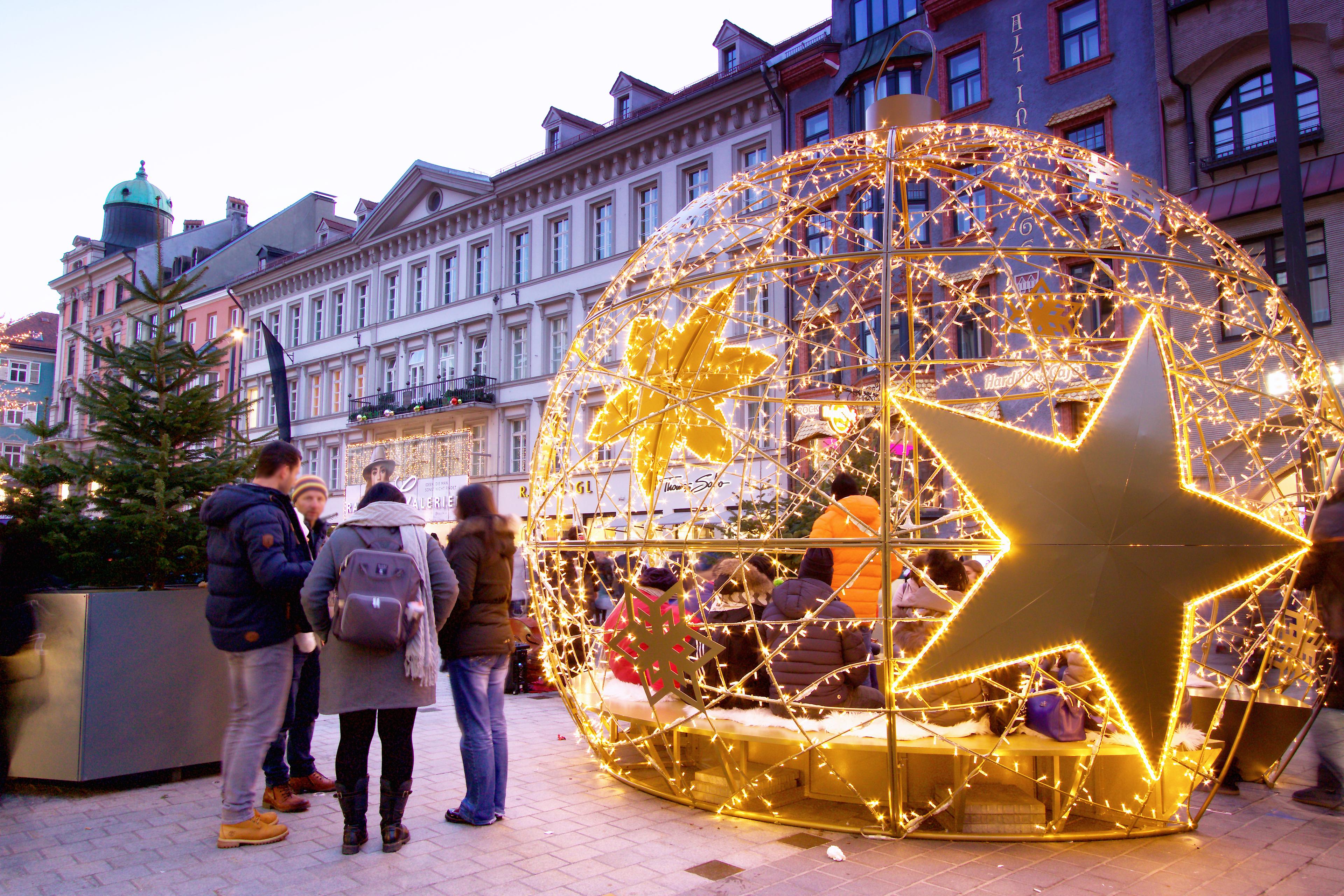 Menschen versammeln sich um eine leuchtende, kugelförmige Weihnachtsdekoration mit Sternen, auf einem städtischen Platz.